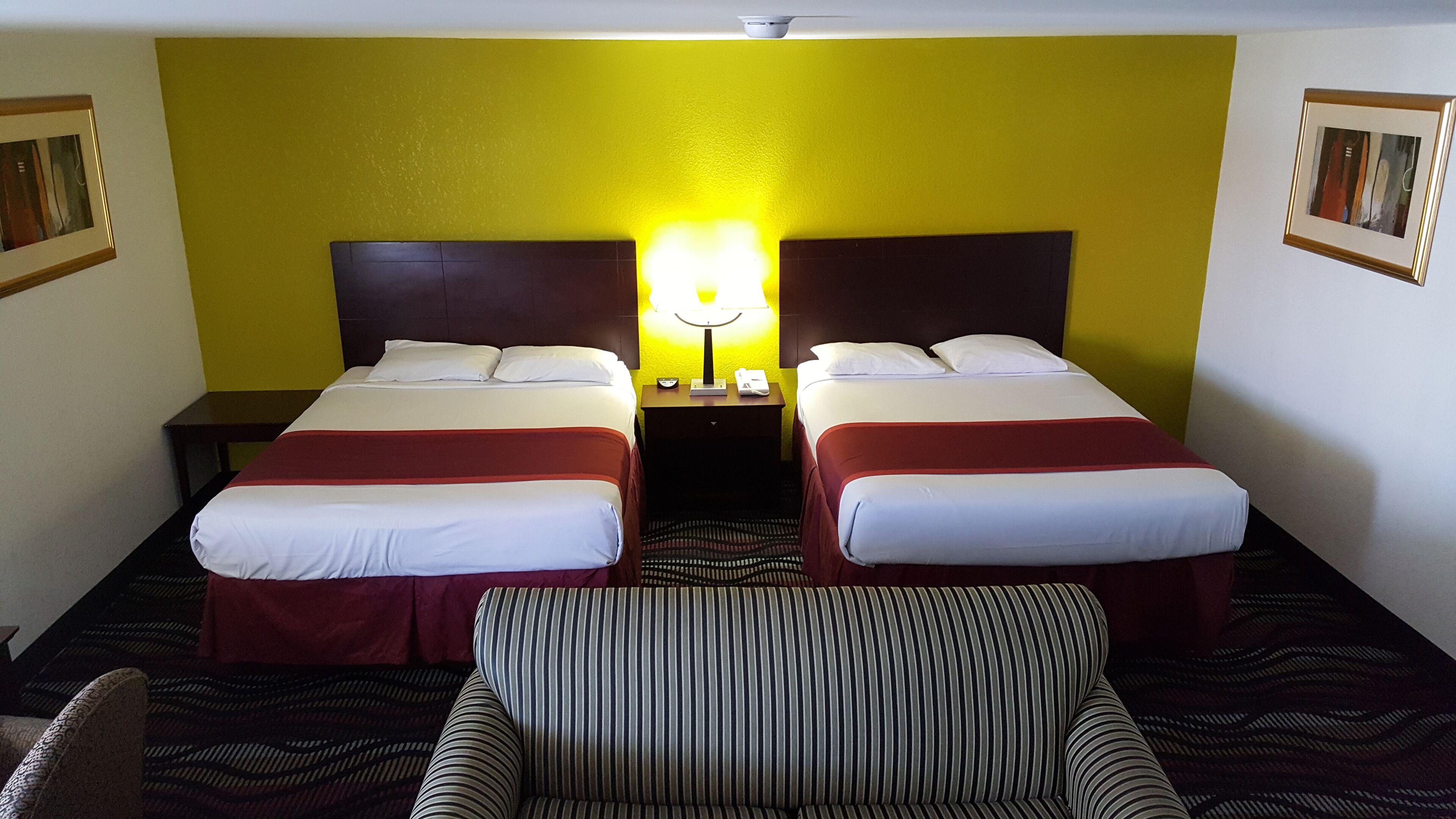 Ein Hotelzimmer mit zwei Betten, einem Nachttisch mit Lampe, einem gestreiften Sessel und einem gemusterten Teppich.
