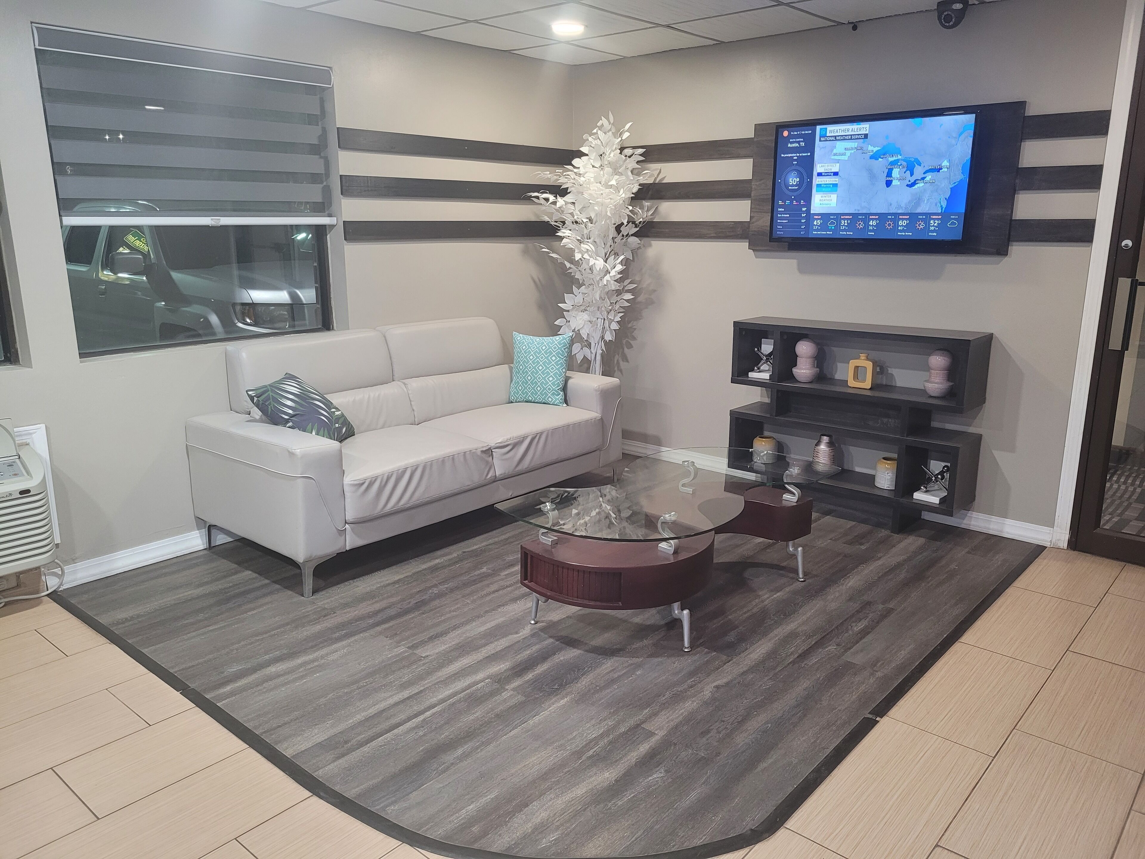 A modern waiting area with a sofa, a glass coffee table, and a mounted TV displaying weather alerts.