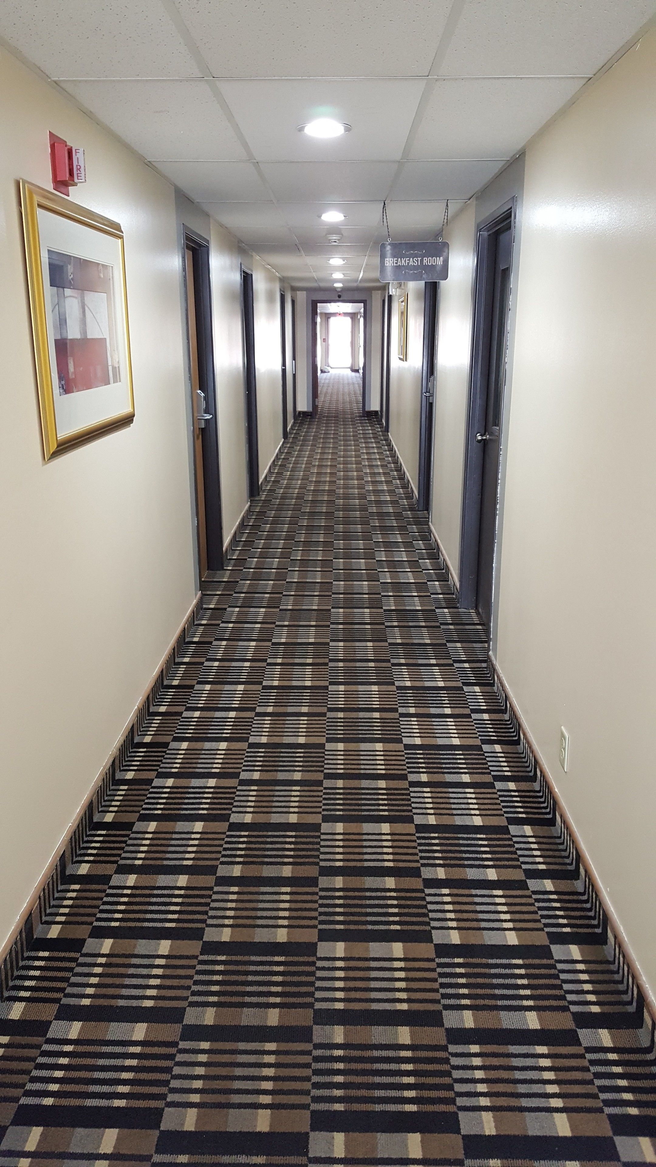 A hotel hallway with a checkered carpet, doors on both sides, and a sign indicating the breakfast room.
