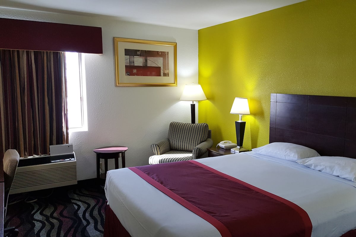 A hotel room with a large bed, two bedside lamps, a striped armchair, a small table, a telephone, and a window with curtains.