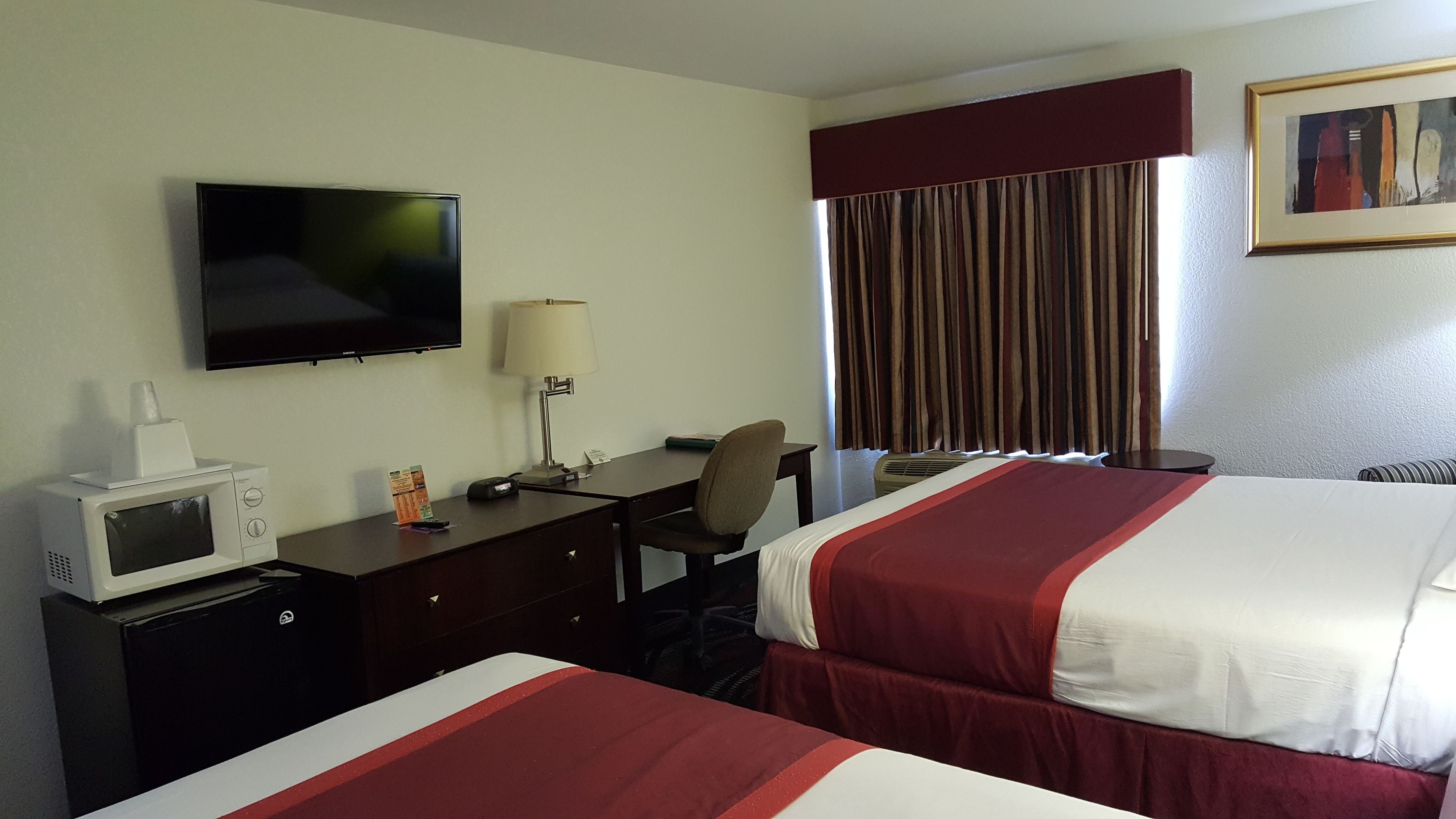 Habitación de hotel con dos camas, un escritorio con computadora, televisor, microondas, lámpara y ventana con cortinas.