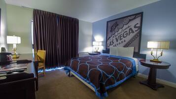 A hotel room with a king-sized bed, a desk with a chair, a table with a lamp, and a wall sign reading "Welcome to Fabulous Las Vegas, Nevada."