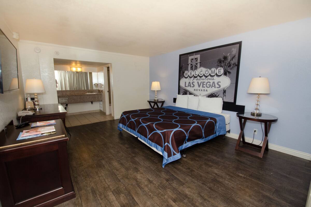A hotel room with a bed, two bedside tables with lamps, a desk with a lamp, a mirror, and a wall with a 'Welcome to Fabulous Las Vegas' sign.