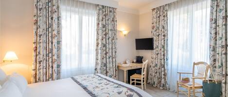 Camera d'albergo con un letto, una scrivania, una sedia, una televisione e tende con motivo naturale.