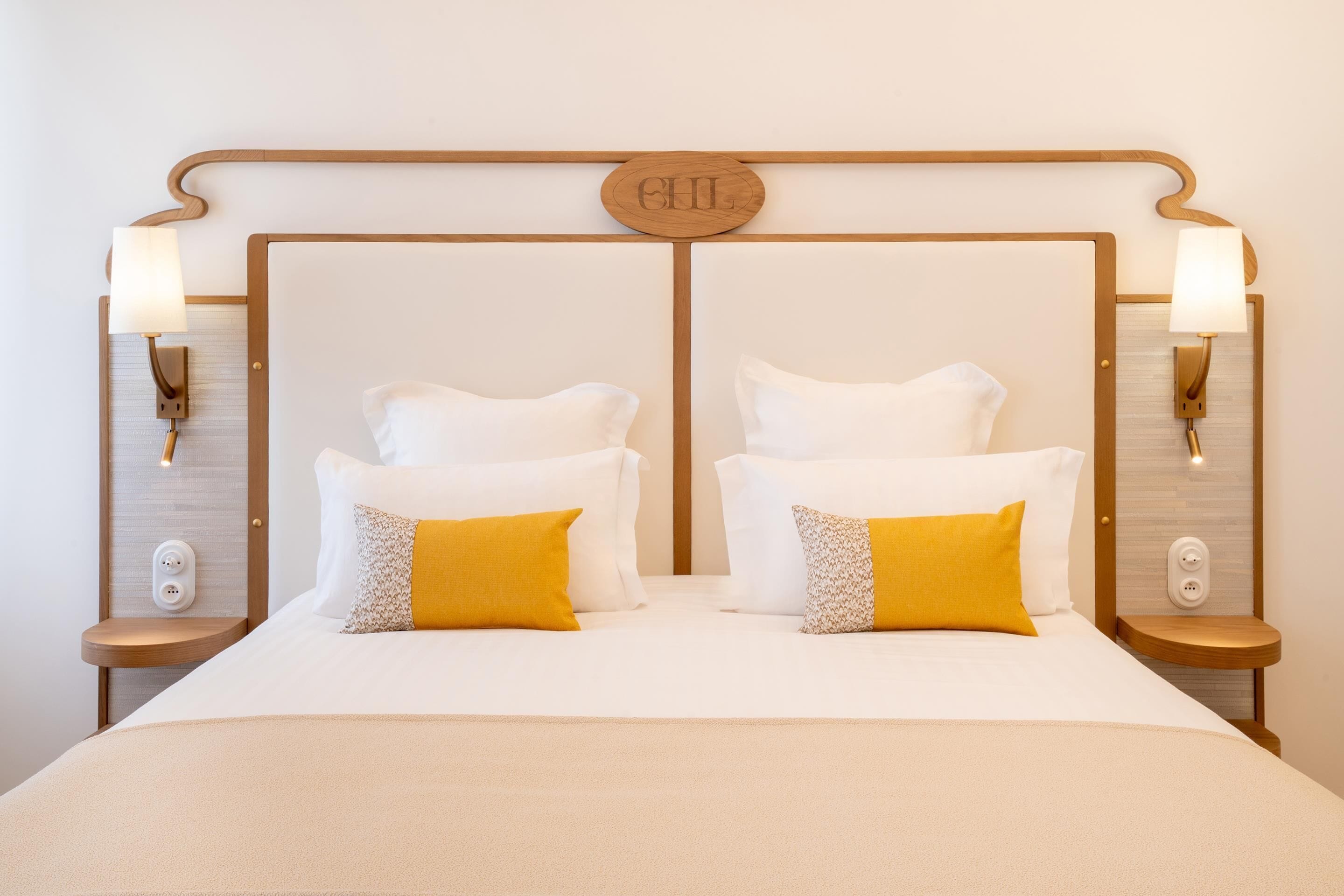 Ein Doppelbett mit zwei weißen und zwei senfgelben Kissen, an dessen Seiten Wandleuchten angebracht sind und das ein Holz-Kopfteil mit einem zentralen Wappen hat.