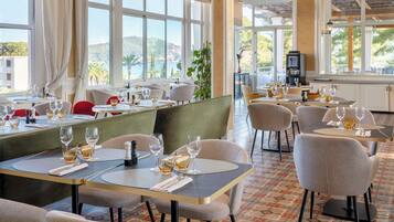 A restaurant with multiple tables and chairs, large windows with a view of the sea, and a patterned floor.