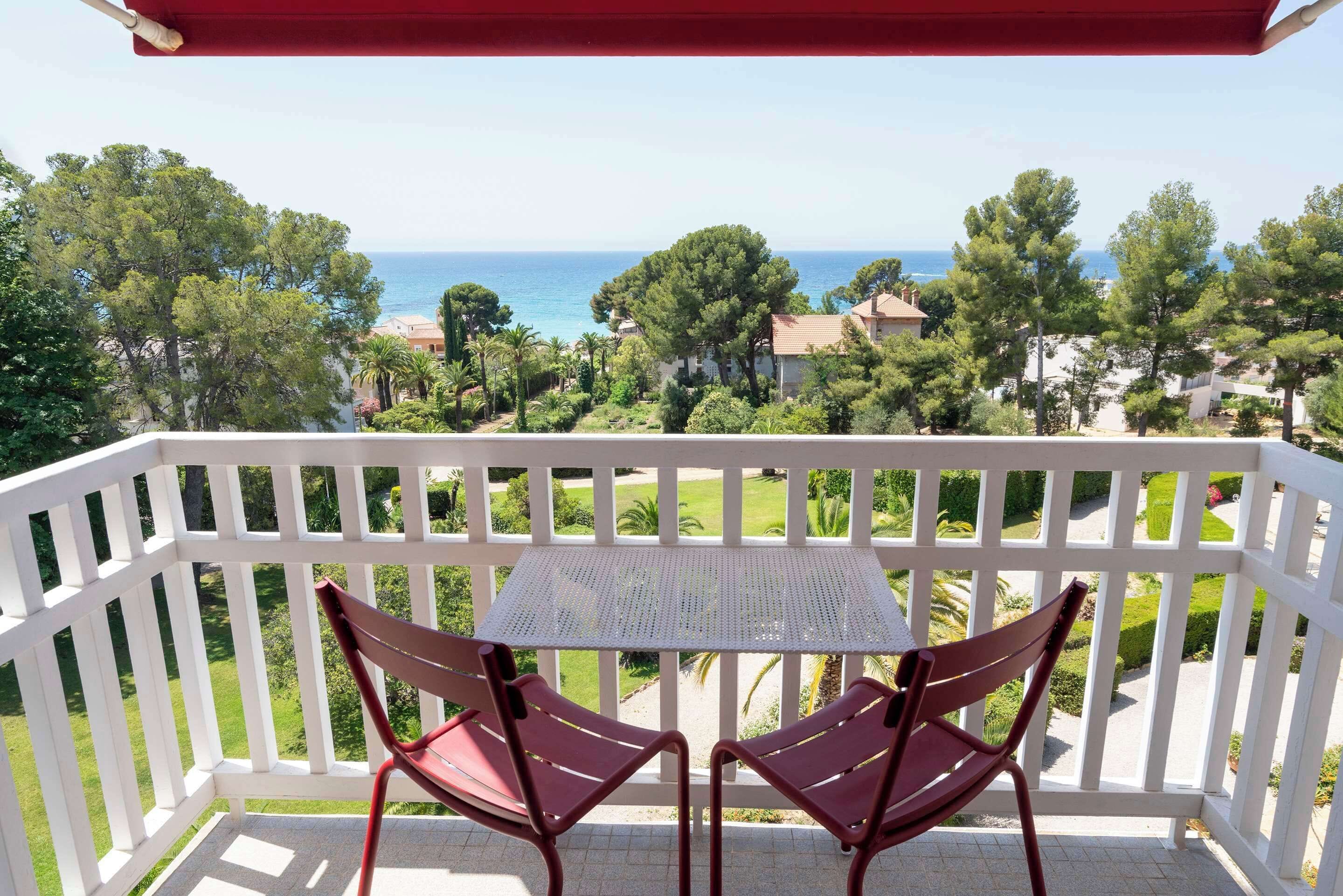 Un balcon avec deux chaises rouges et une petite table, donnant sur une zone côtière bordée d’arbres et de maisons.