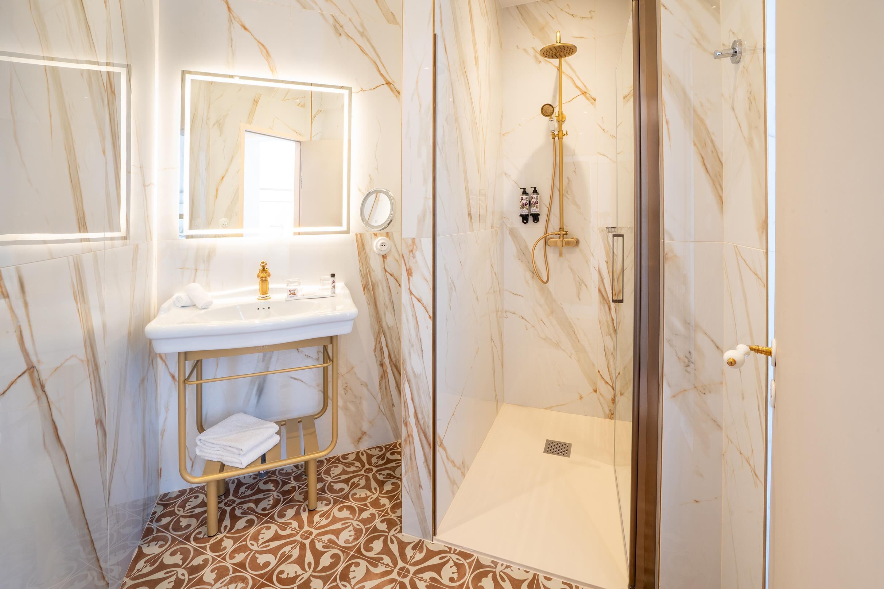 A bathroom with a marble wall, a gold vanity, a shower with a gold showerhead, and a patterned floor.