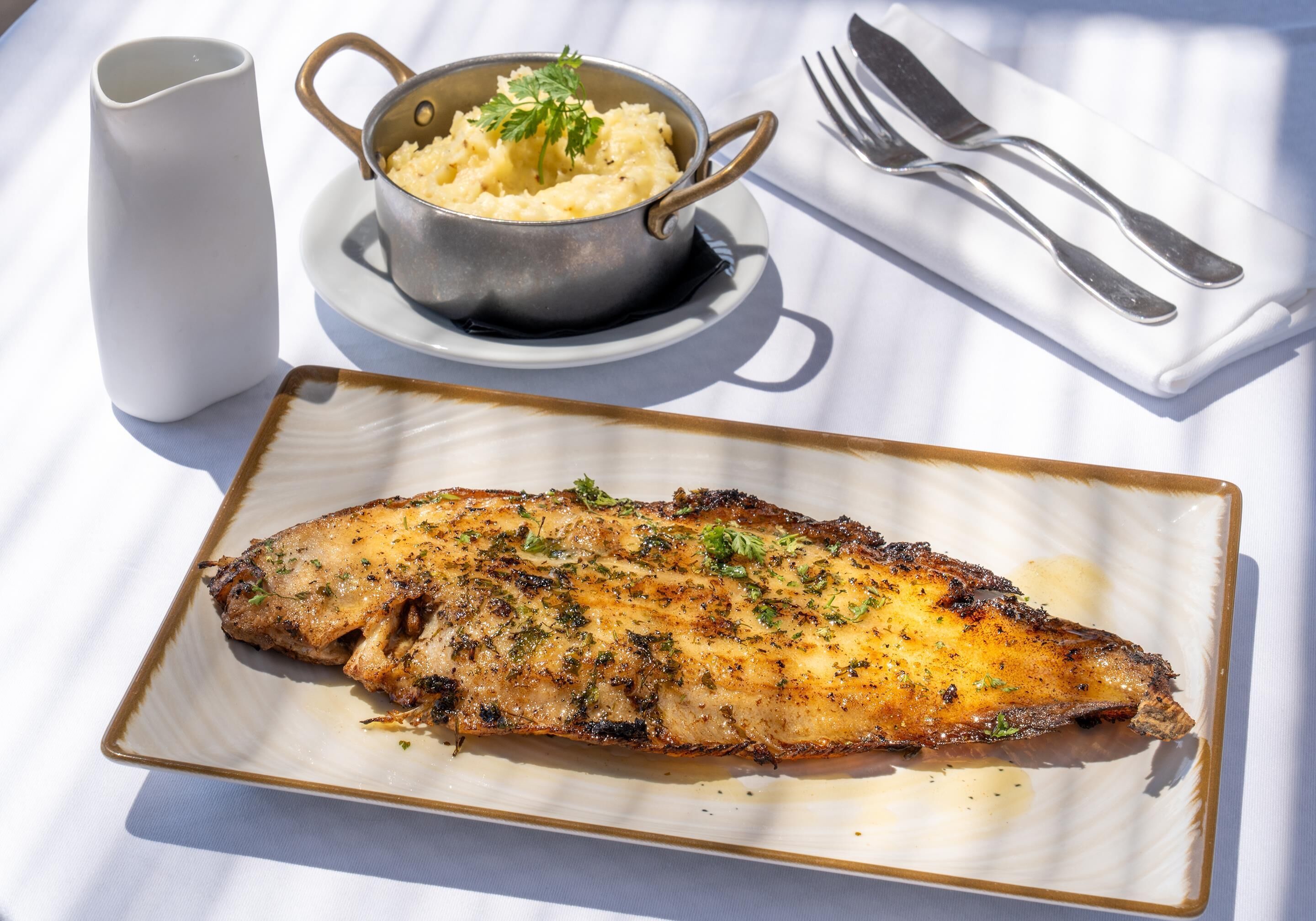 Un plato de pescado a la parrilla con hierbas, un bol de puré de papas y una taza blanca sobre una mesa con servilleta y cubiertos blancos.