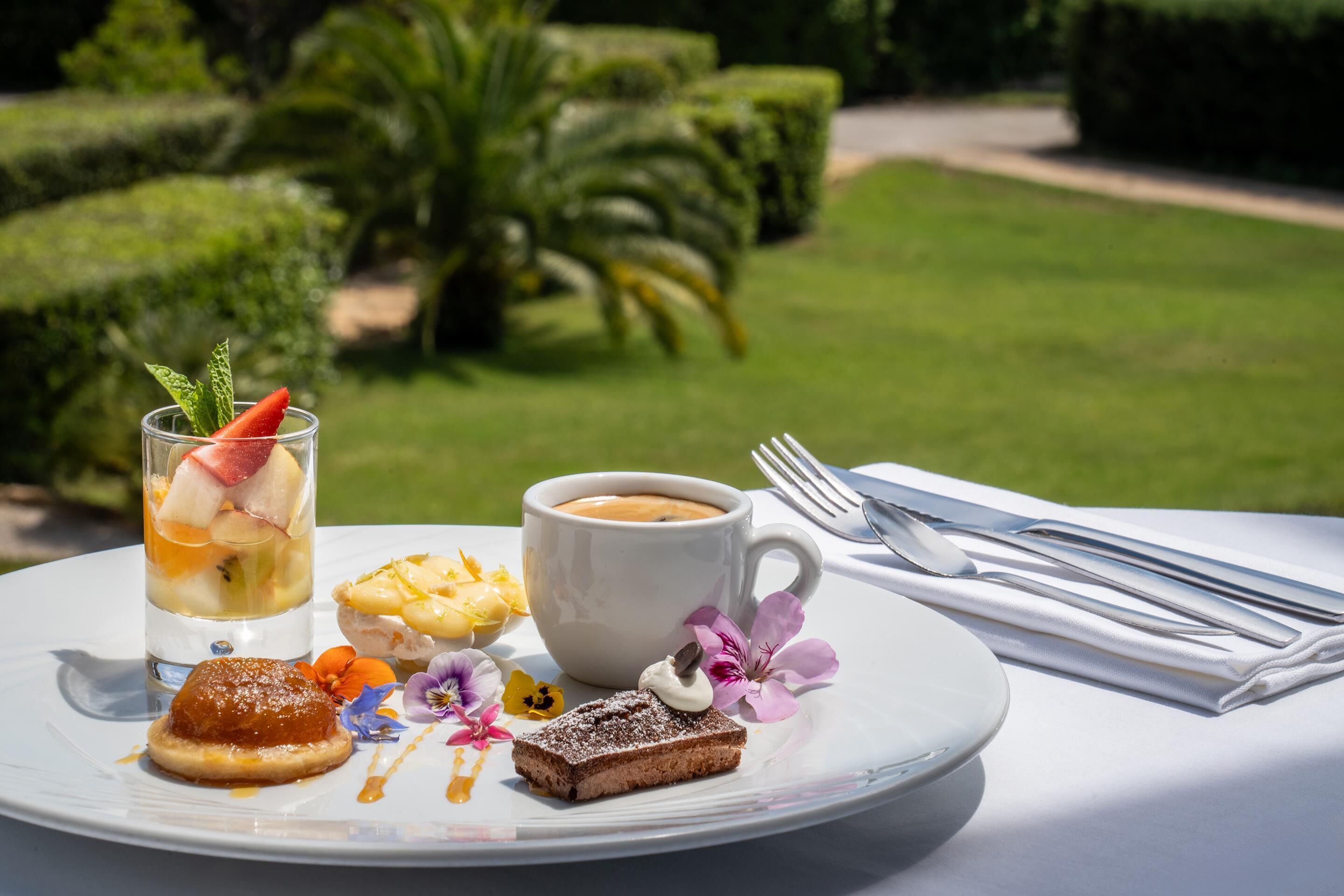 Un plato con postre, una taza de café y un vaso de jugo de frutas sobre una mesa al aire libre.