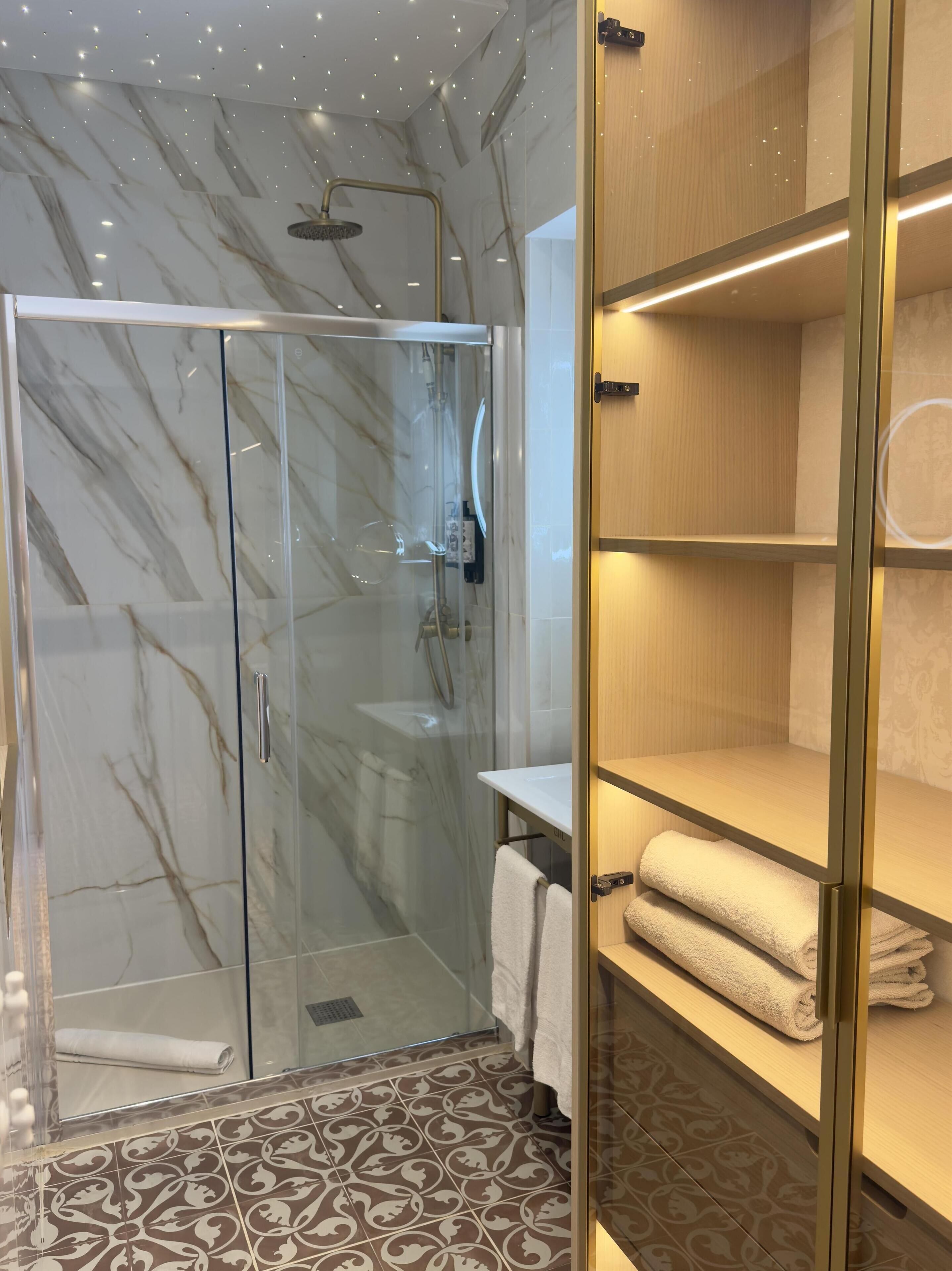 A modern bathroom with a glass-enclosed shower, marble walls, patterned floor tiles, and wooden shelves with towels.