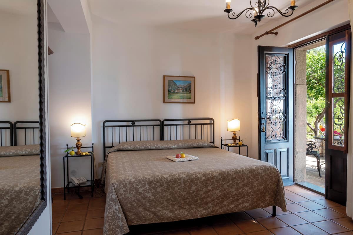 Une chambre d’hôtel avec un lit, deux tables de chevet avec des lampes, une petite table avec des fruits, un tableau au mur et une porte donnant sur un espace extérieur.