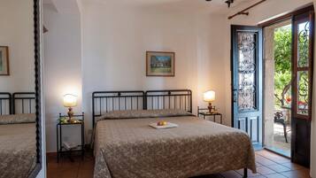 A hotel room with a bed, two bedside tables with lamps, a small table with fruit, a painting on the wall, and a door leading to an outdoor area.