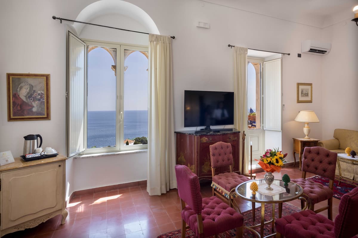 Un salon avec un téléviseur, une table à manger avec des chaises, un canapé et une vue sur la mer grâce à de grandes fenêtres.