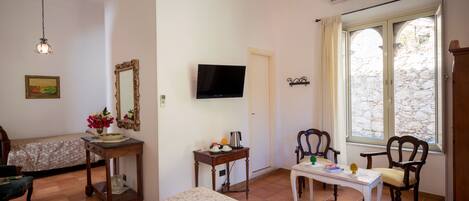 A room with a bed, a table with a chair, a television, a mirror, and a window with a stone wall view.