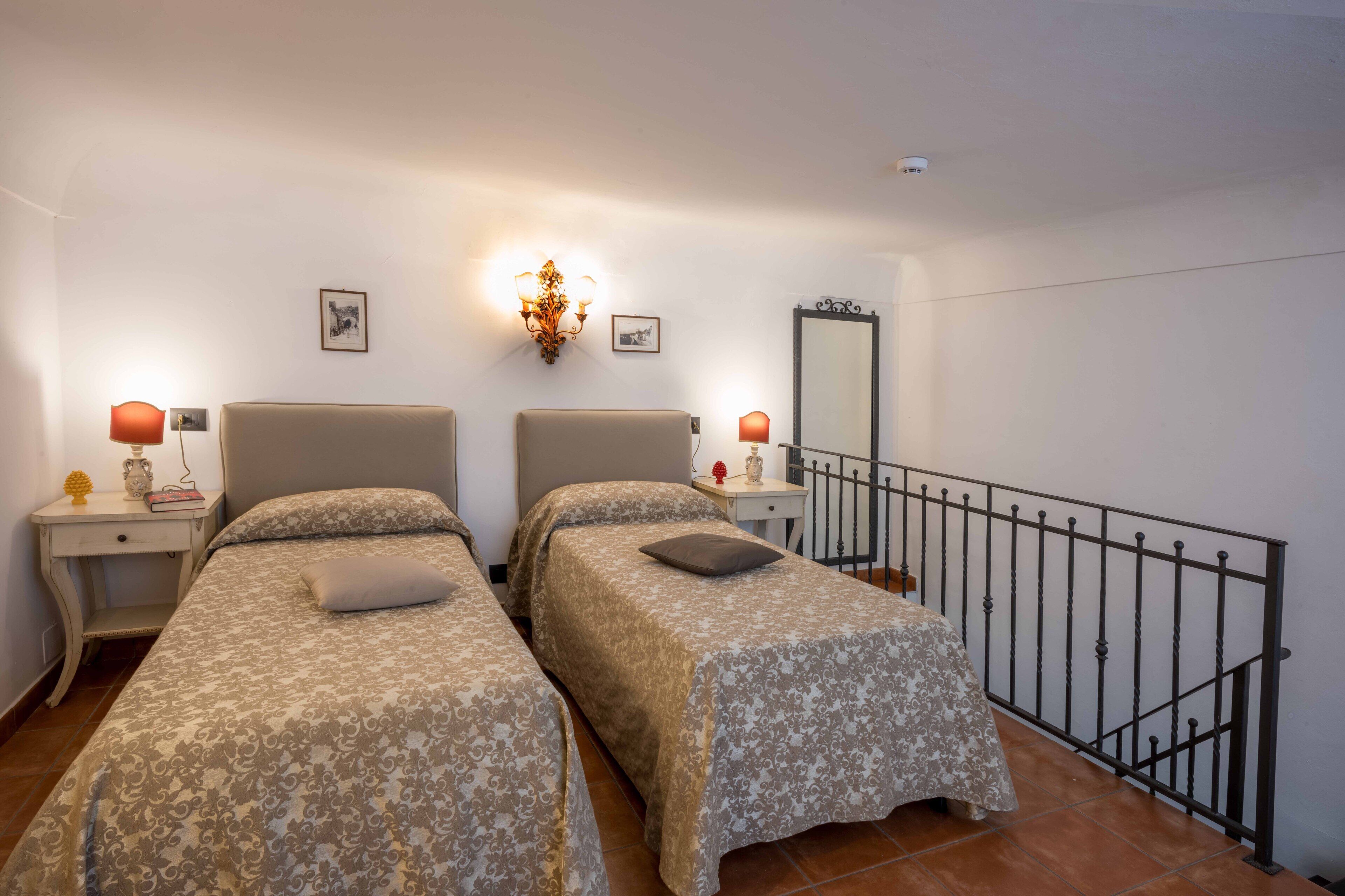 A room with two beds, a nightstand with a lamp, a wall-mounted light fixture, a mirror, and a balcony railing.