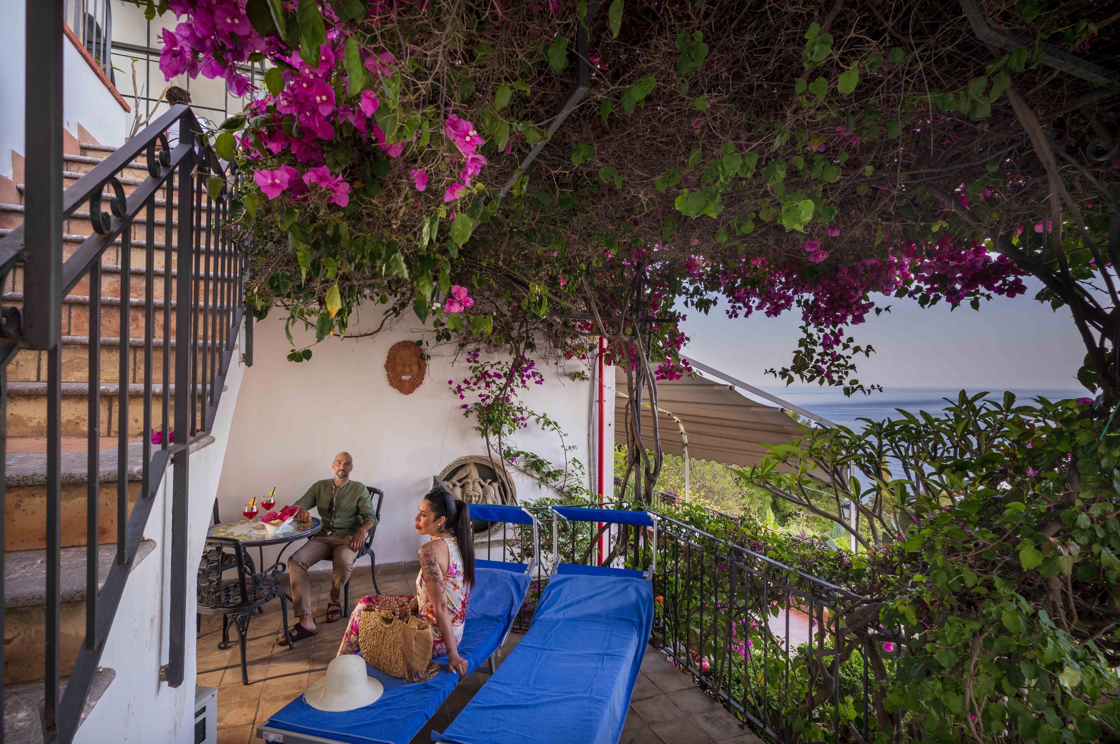 Un balcone con un divano blu, un tavolino con dei drink e una persona seduta.