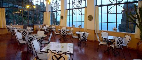 Un restaurante con varias mesas y sillas, ventanas amplias con celosías de hierro decorativas, y vistas al exterior.