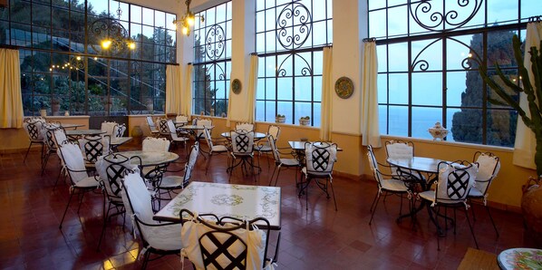 A restaurant with multiple tables and chairs, large windows with decorative iron grilles, and a view of the outdoors.