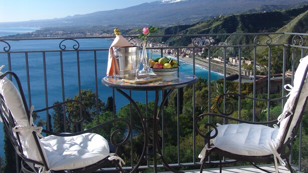 Un balcon avec deux fauteuils blancs dotés de coussins, une table en métal sur laquelle se trouvent une bouteille, un verre et un saladier rempli de fruits, offrant une vue sur la côte et, au loin, sur une montagne dont le sommet est enneigé.