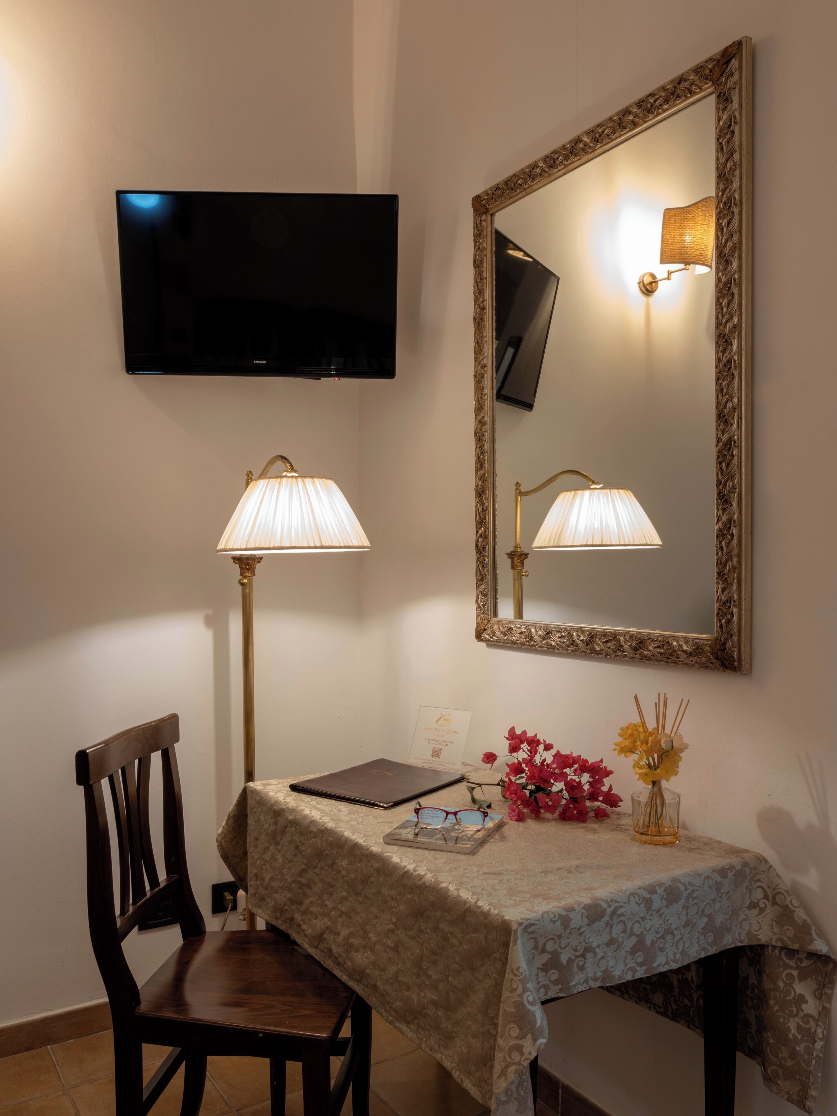 Una habitación con una televisión instalada en la pared, un espejo grande y ornamentado, dos lámparas fijadas a la pared, una silla de madera, una mesa con mantel estampado, una computadora portátil, un florero con flores y un pequeño adorno.