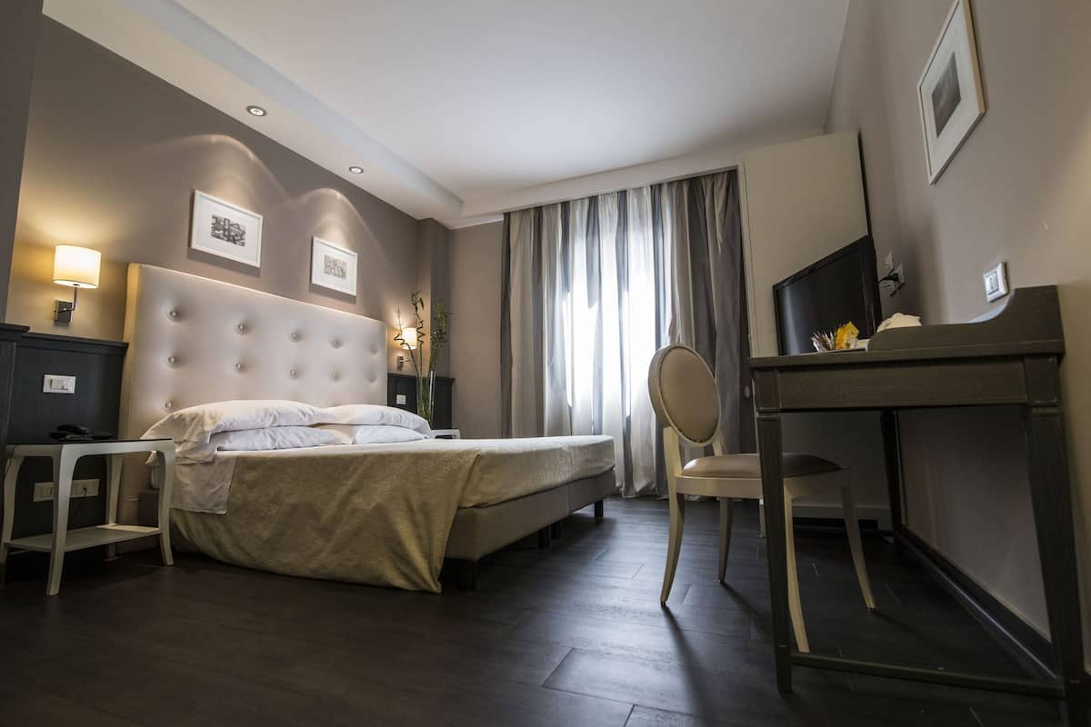 A hotel room with a large bed, a desk, a chair, a TV, and a lamp.