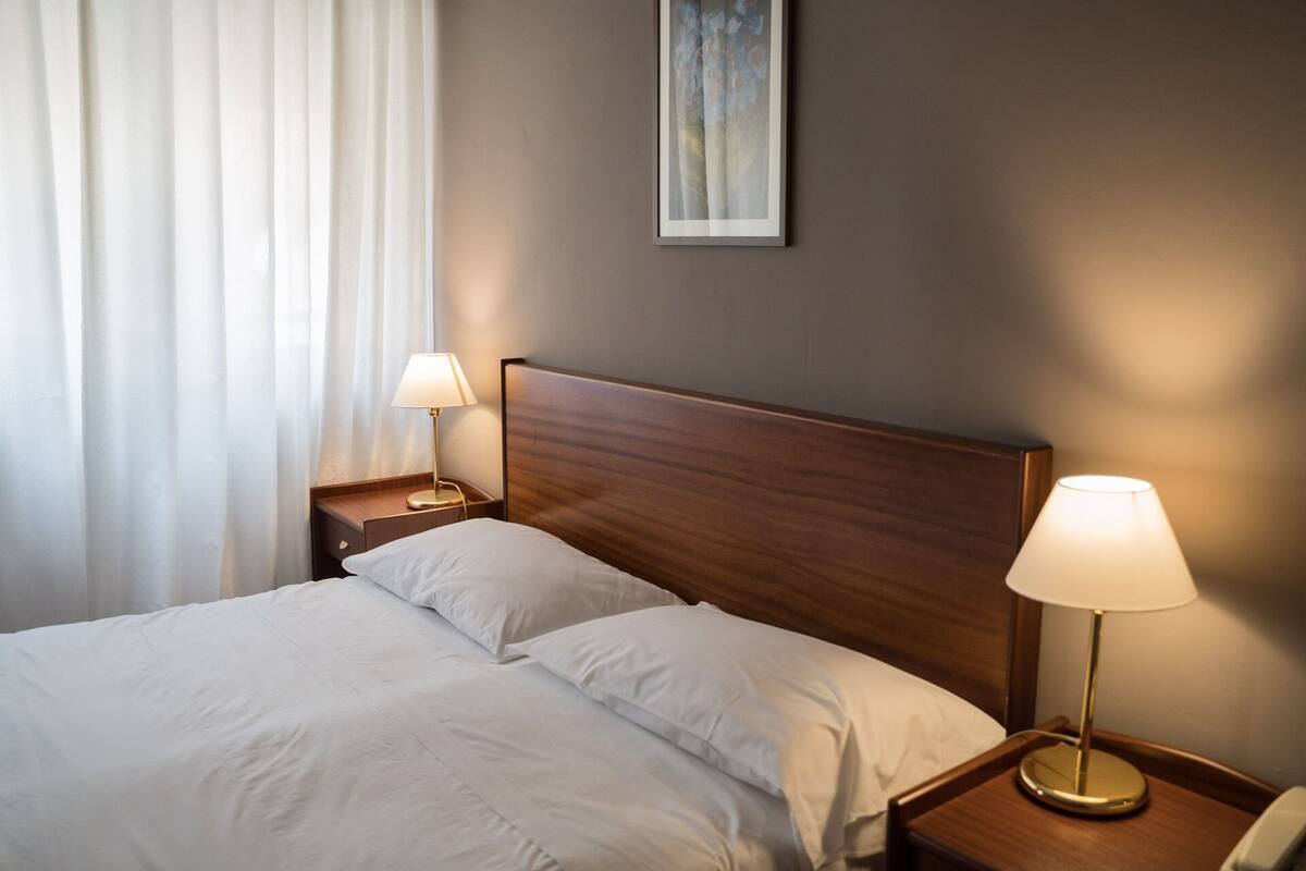 A neatly made bed with white linens, two bedside lamps, and a wooden headboard.