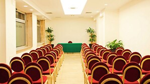 Un salón de conferencias con filas de sillas rojas, una mesa verde y plantas en macetas.