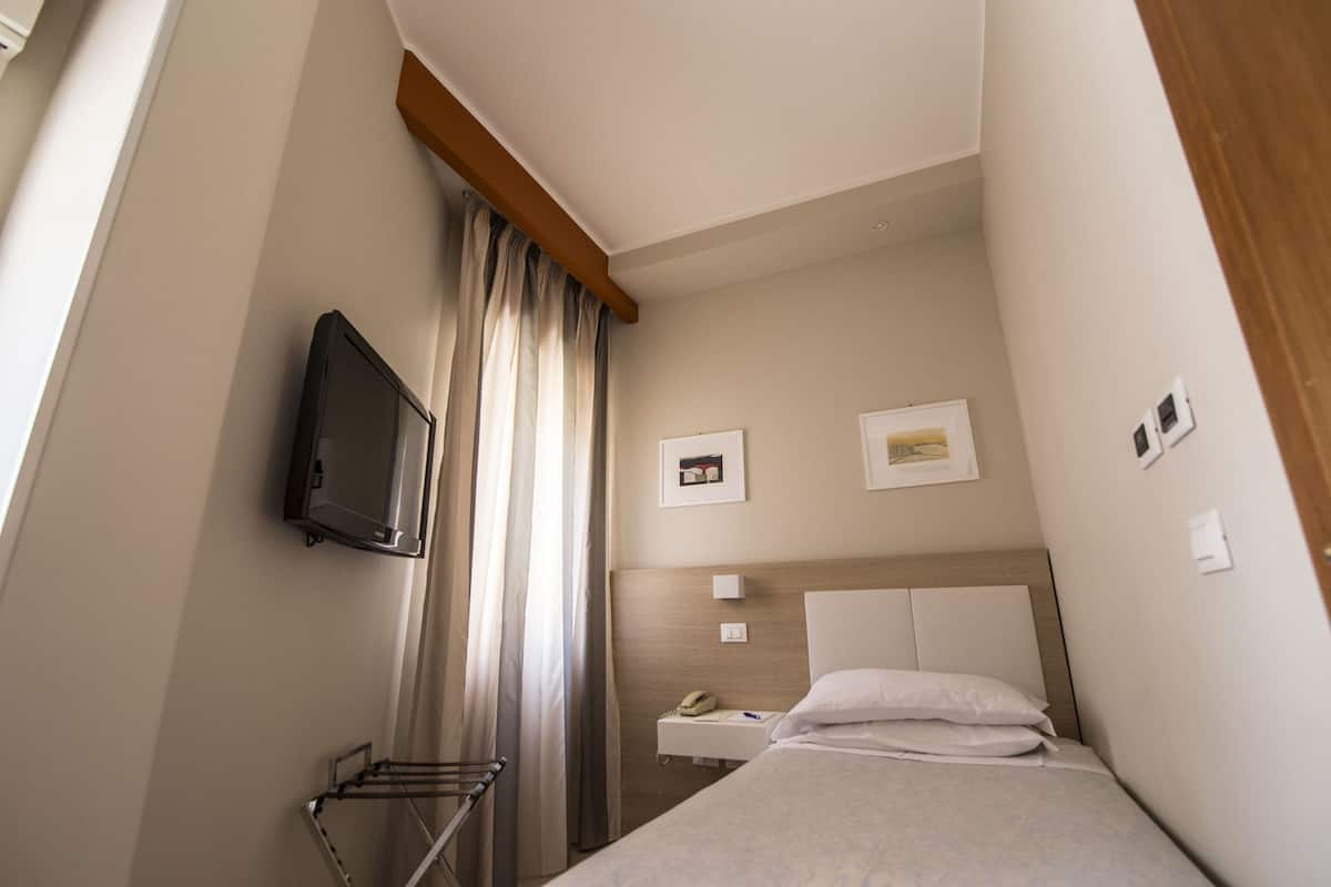 A hotel room with a bed, a TV mounted on the wall, a small bedside table with a phone, and a window with curtains.