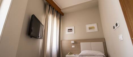 A hotel room with a bed, a TV mounted on the wall, a small bedside table with a phone, and a window with curtains.