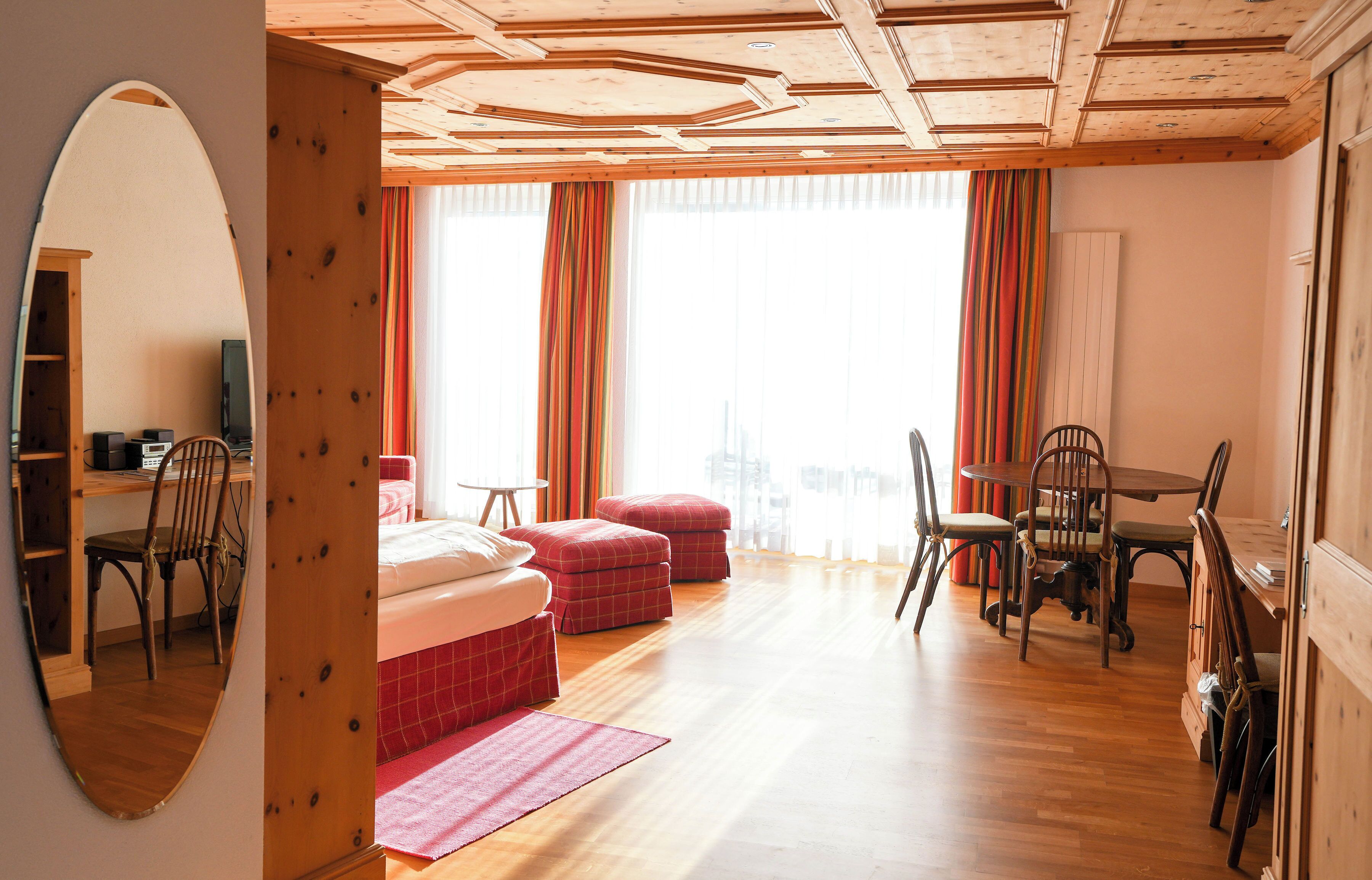 A room with a wooden ceiling, a large mirror, a bed, a desk, a chair, a table, and a window with curtains.
