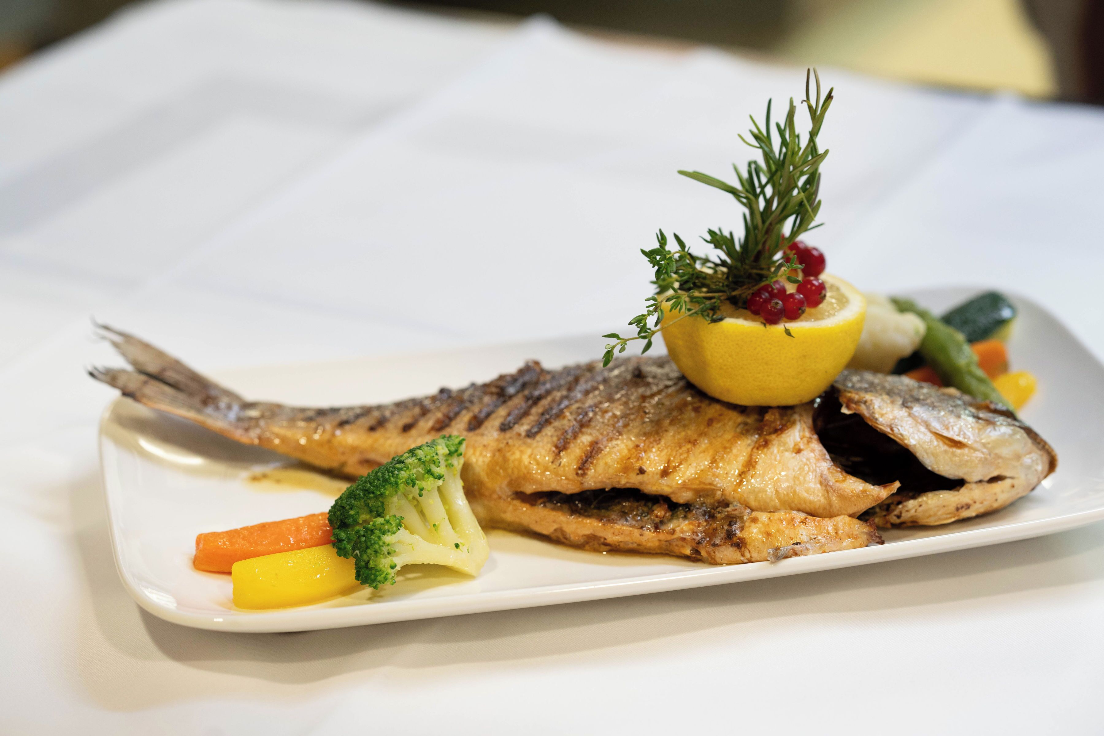 A white plate with a grilled fish, broccoli, carrots, and a lemon garnish.
