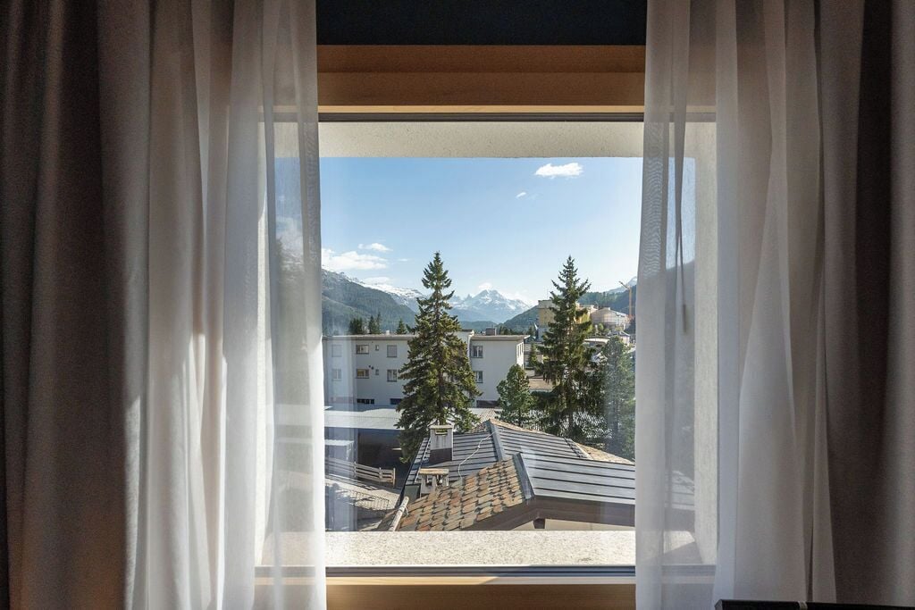 A window with sheer curtains, a view of a building with a sloped roof, and snow-capped mountains in the background.