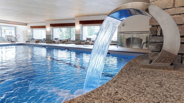 An indoor swimming pool with a waterfall feature, lounge chairs, and a stone wall. - Hotel Bären (St. Moritz)