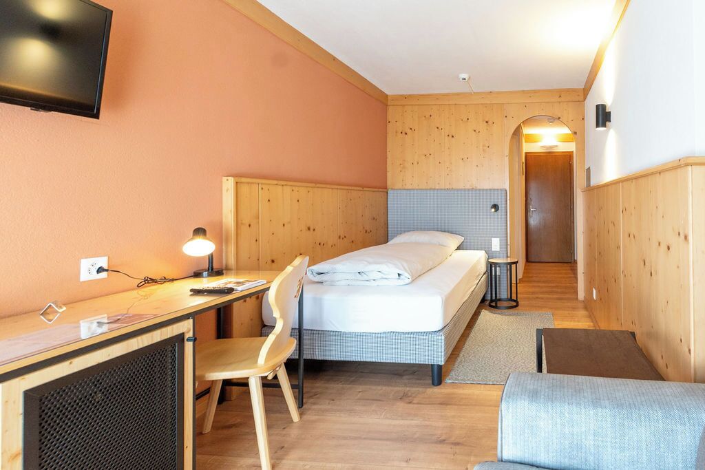 Habitación de hotel pequeña con una cama, un escritorio con una silla, un televisor y una mesita de noche con lámpara.