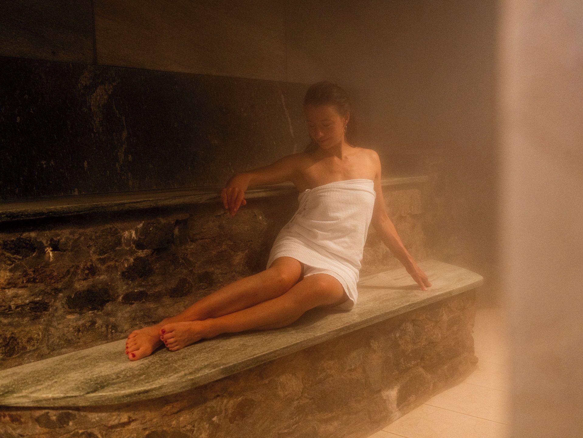 A person sitting on a stone bench in a dimly lit room with a rough wall texture.