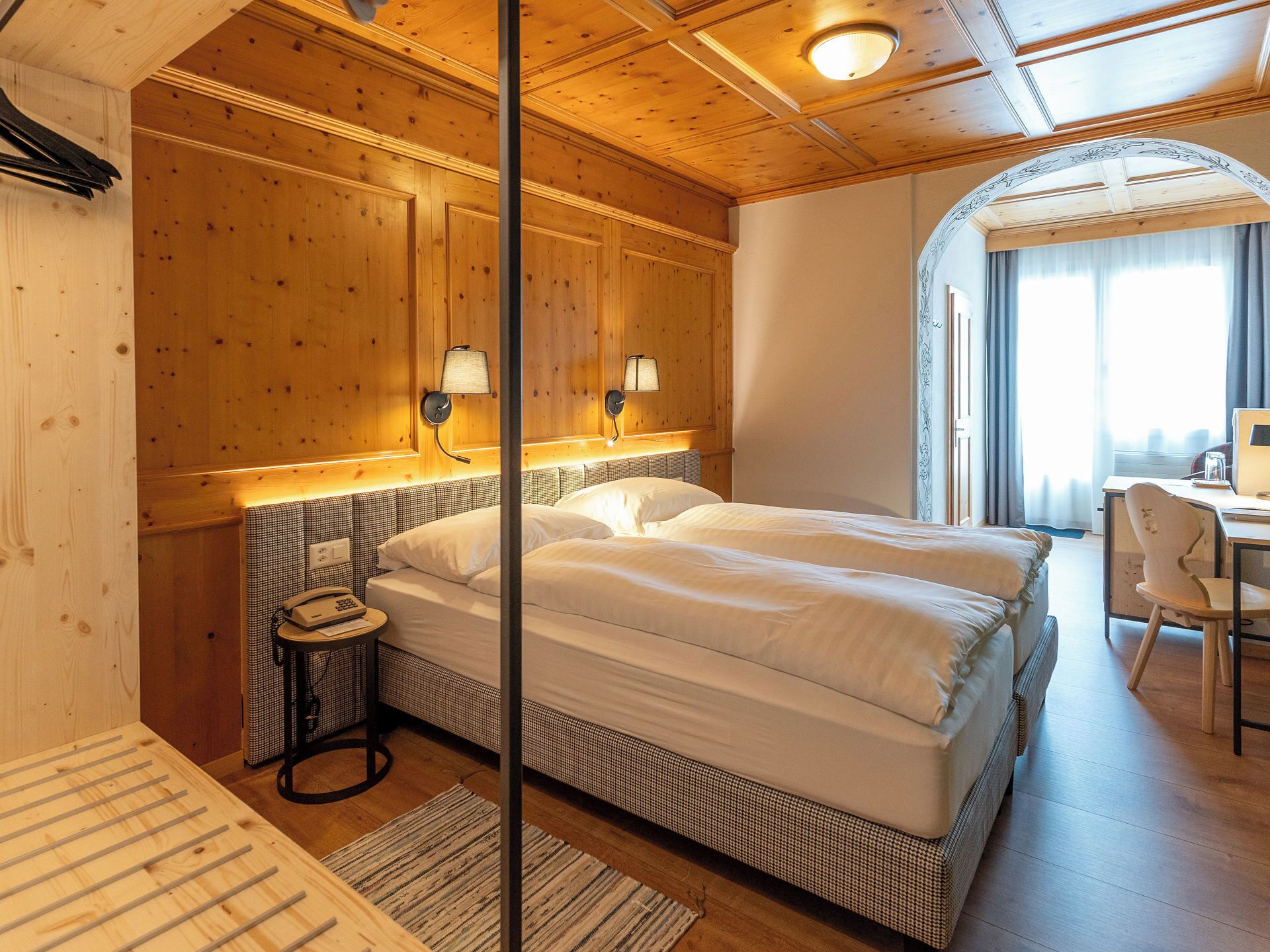 A hotel room with a wooden ceiling, a bed with white bedding, two bedside lamps, a desk with a chair, and a window with curtains.