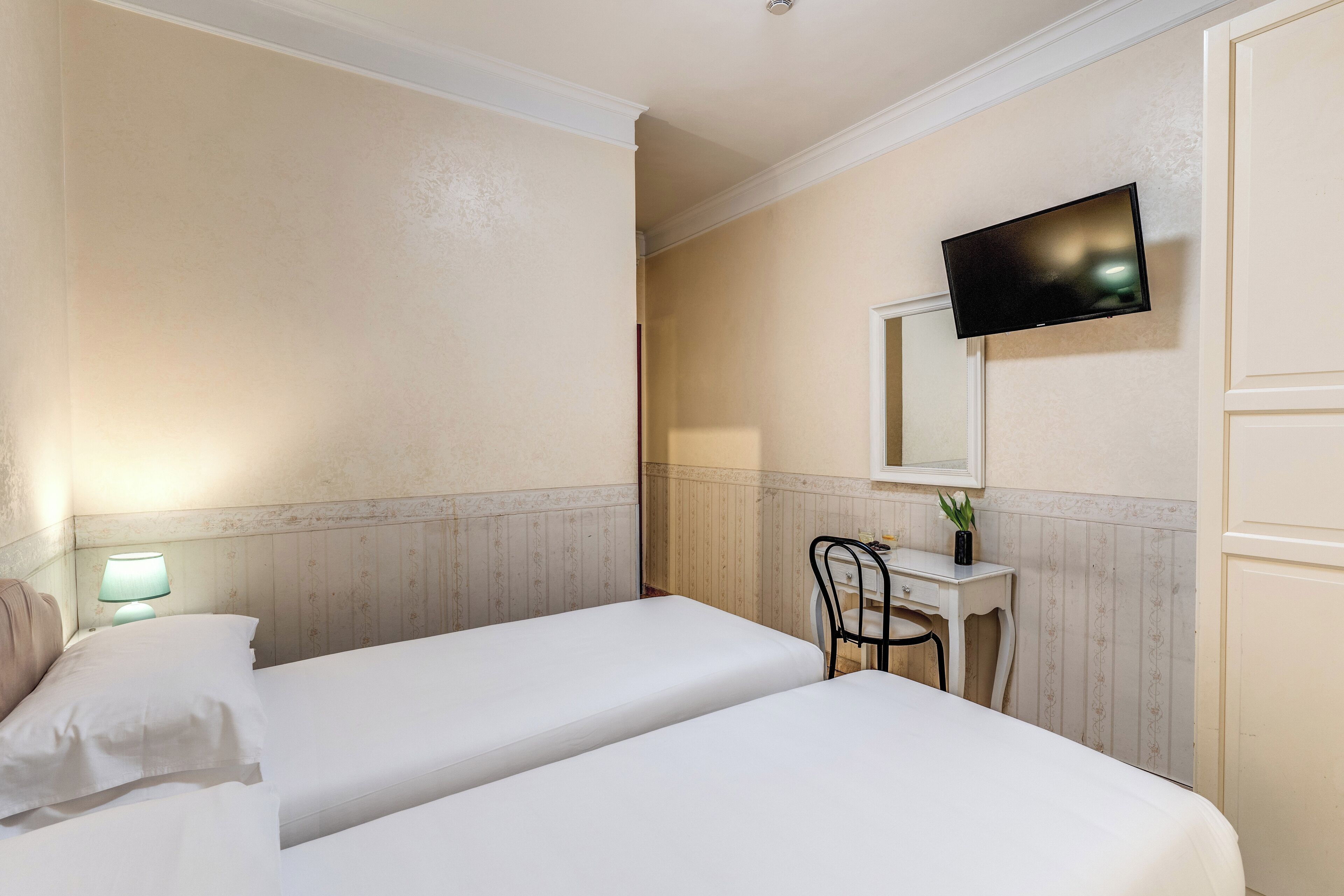 A hotel room with two single beds, a desk with a chair, a television mounted on the wall, and a small table with a plant.