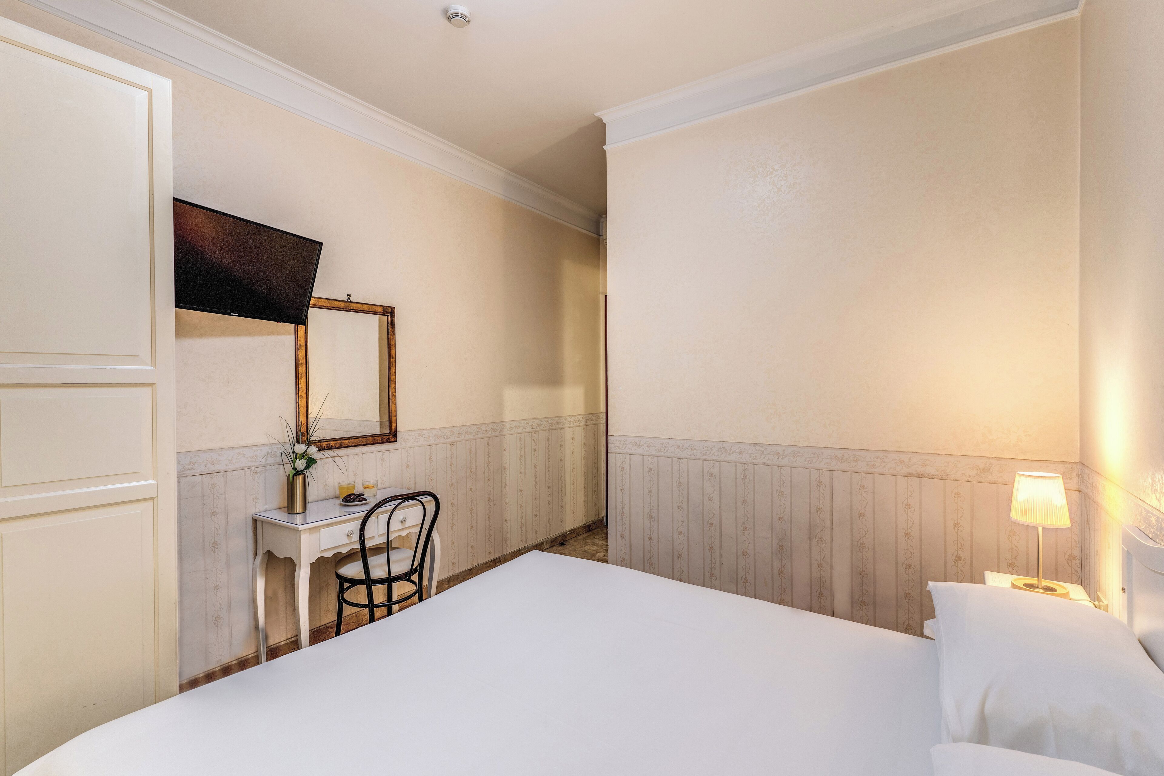 Hotellihuone, jossa on sänky, työpöytä tuolilla, peili ja yöpöytä lampun kanssa.