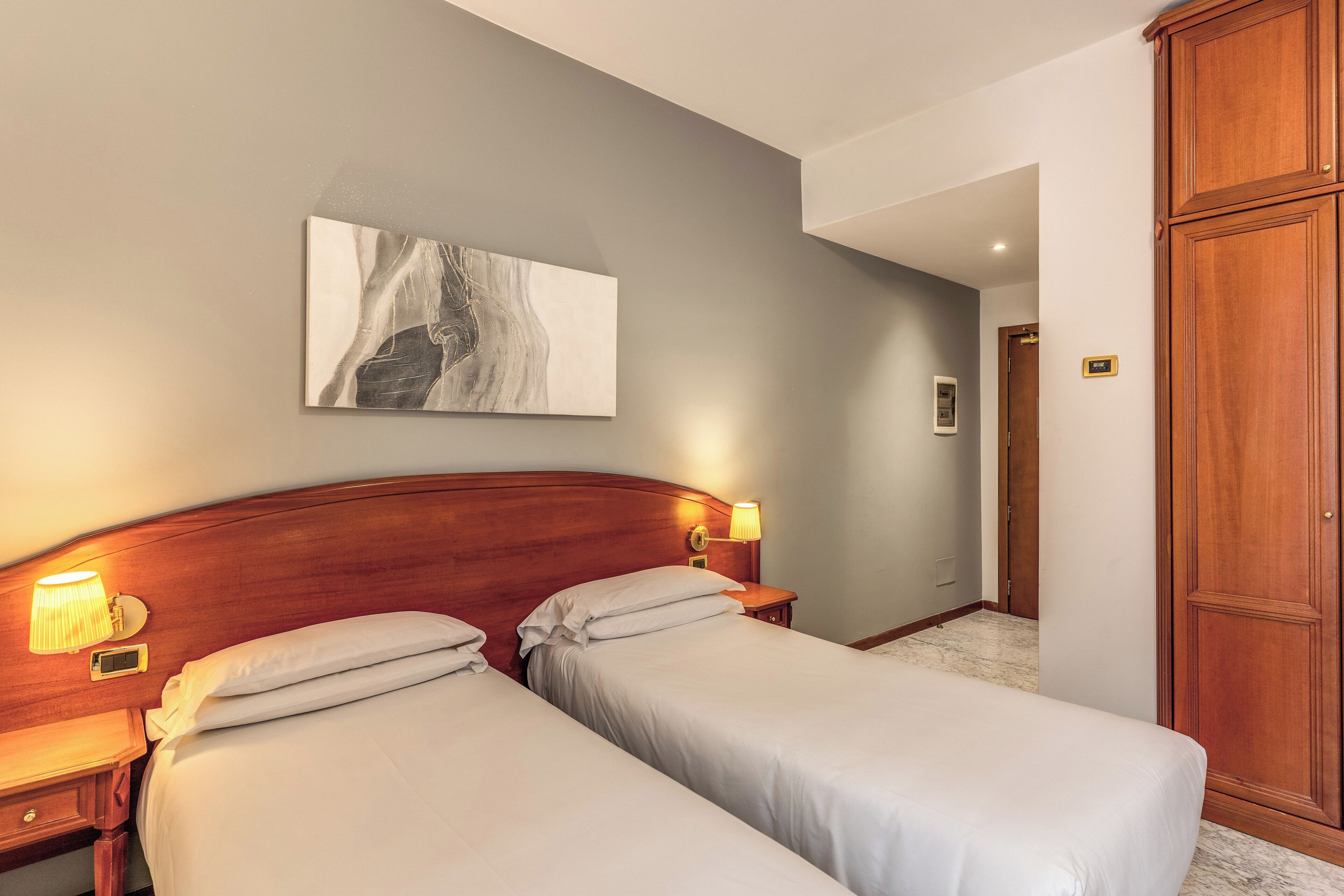 A hotel room with two beds, a wooden headboard, bedside lamps, a marble floor, and a wooden wardrobe.