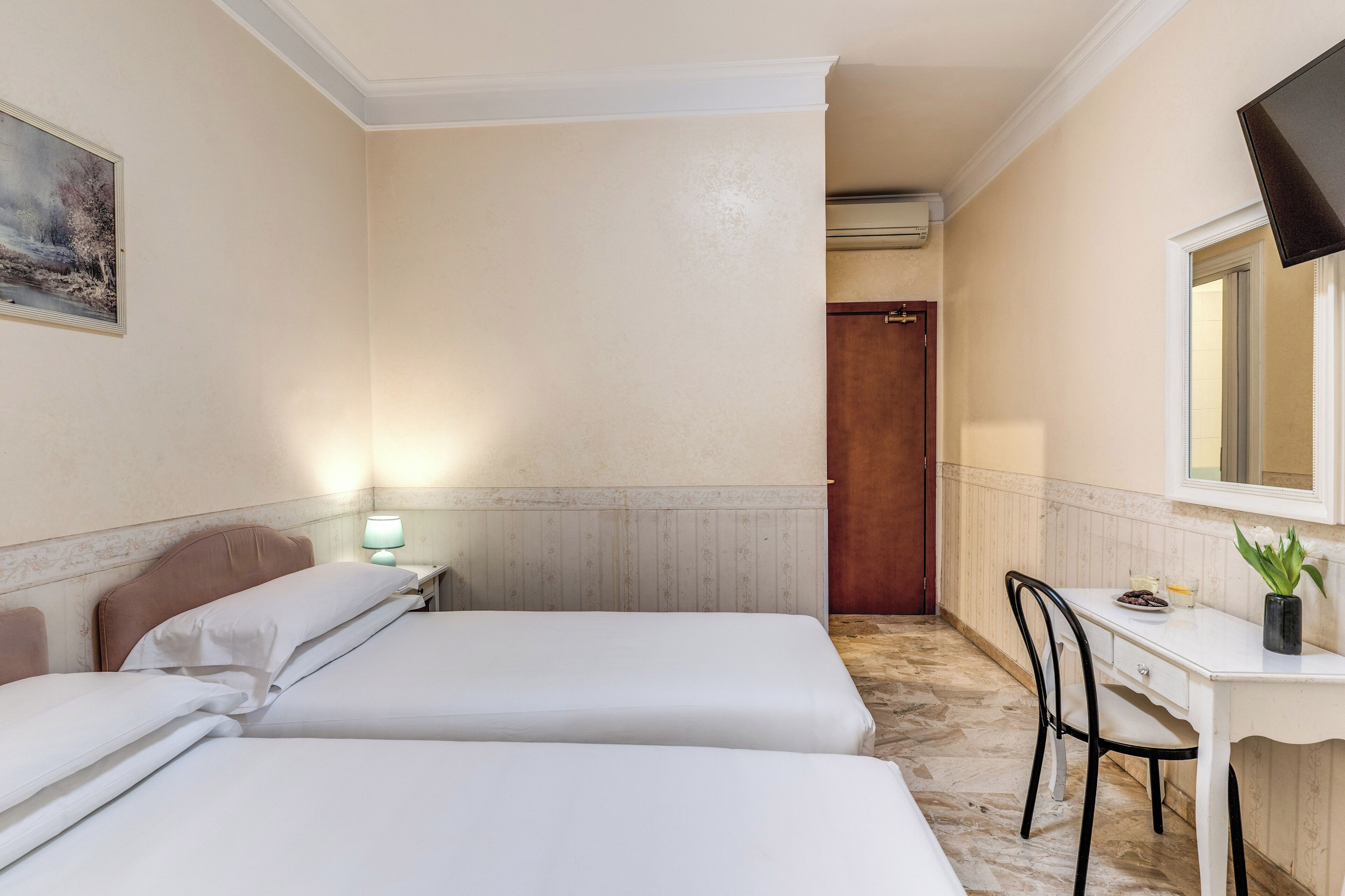 A hotel room with two beds, a desk, a chair, a painting, and a mirror.