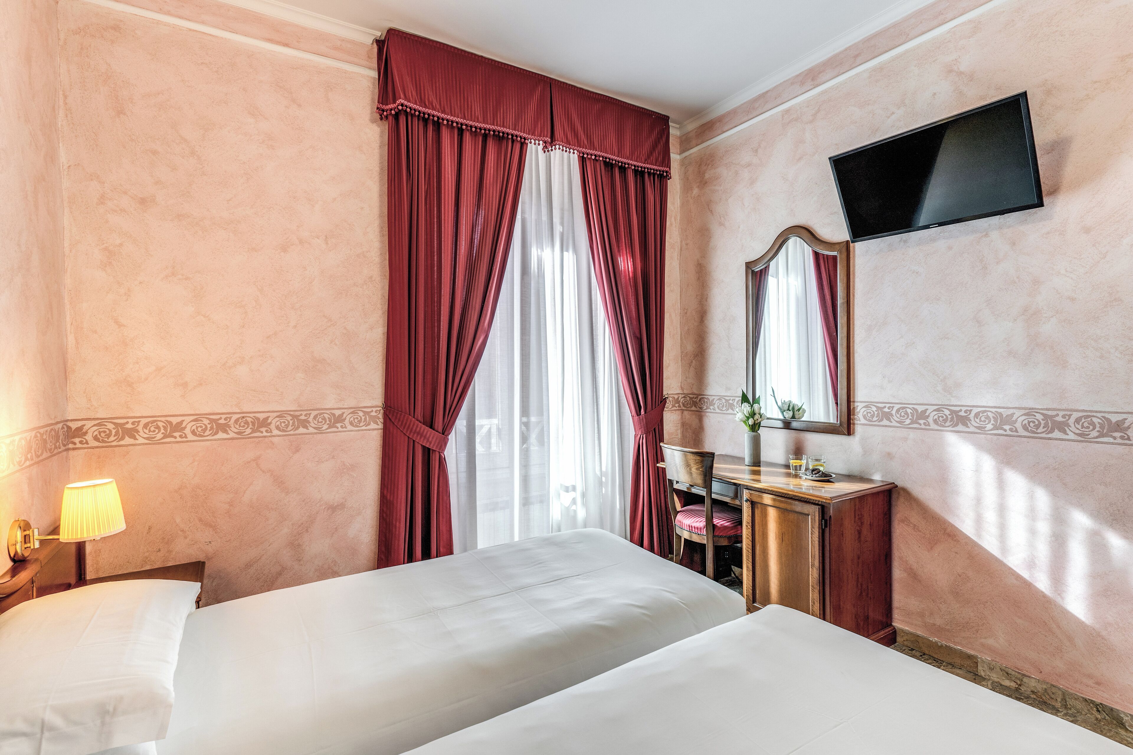 Une chambre d’hôtel avec deux lits, un téléviseur fixé au mur, un bureau avec une chaise et une lampe sur la table de chevet.