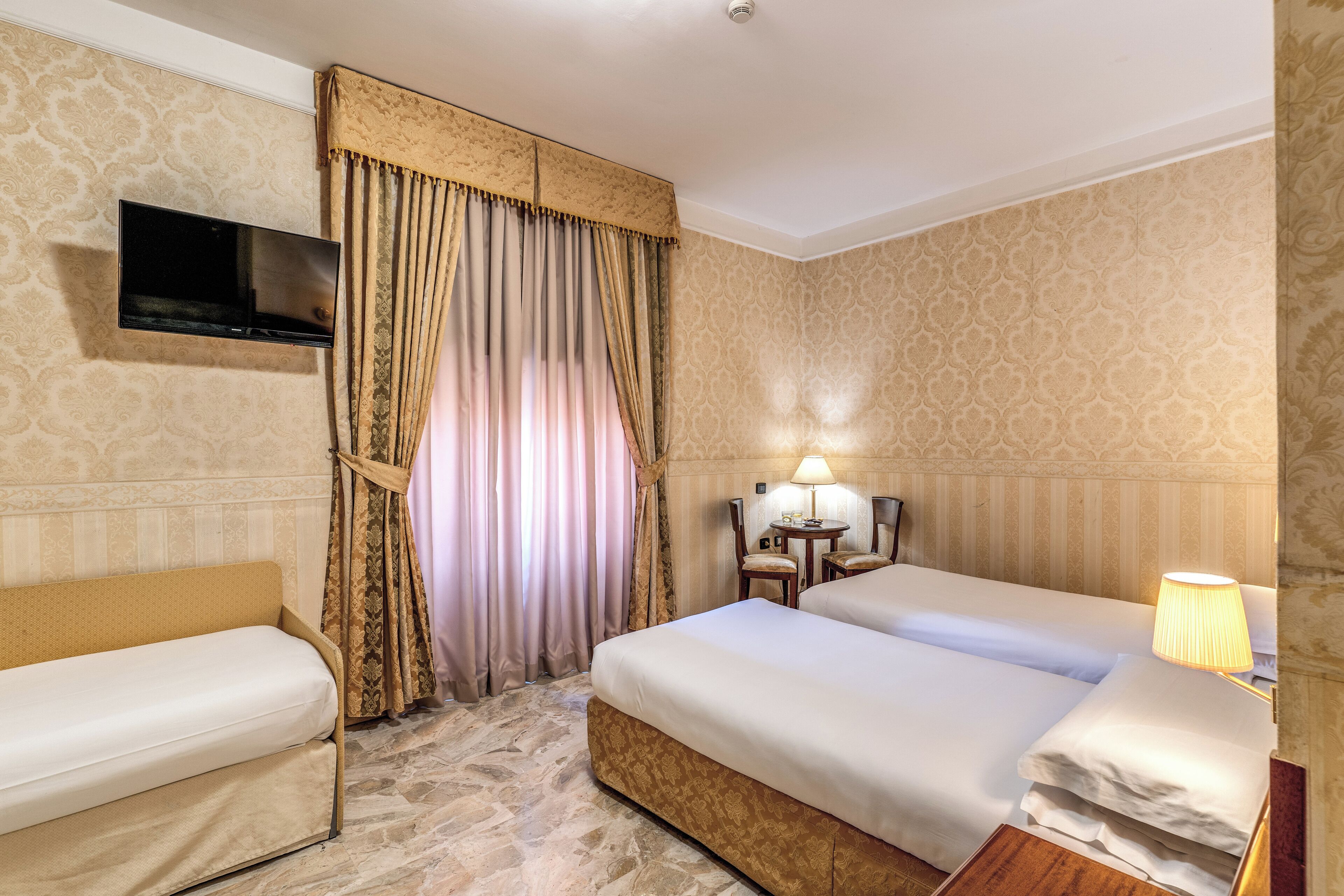 A hotel room with a bed, a sofa, a TV mounted on the wall, a lamp, and curtains.