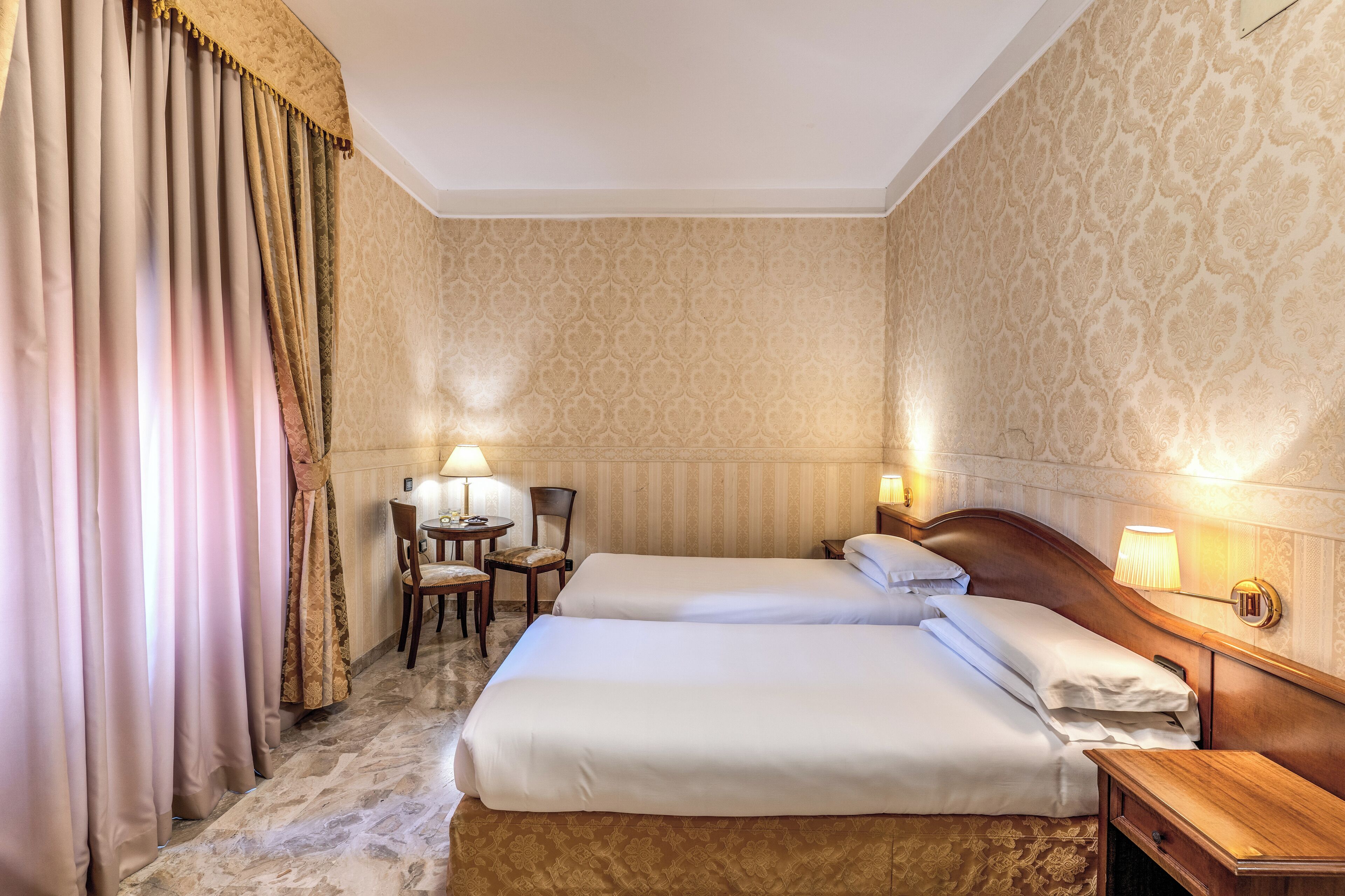 Une chambre d’hôtel avec deux lits, un bureau avec une chaise, une lampe et un papier peint à motifs.