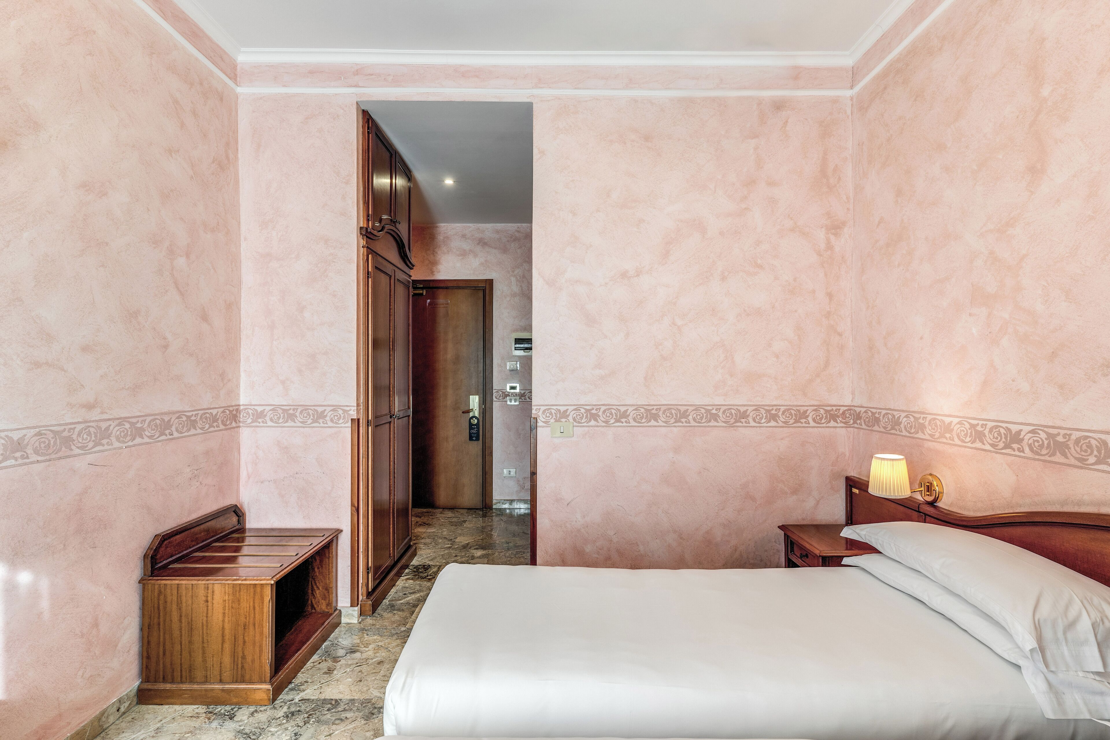 A hotel room with a bed, a nightstand with a lamp, a wooden bench, and a door leading to a hallway.