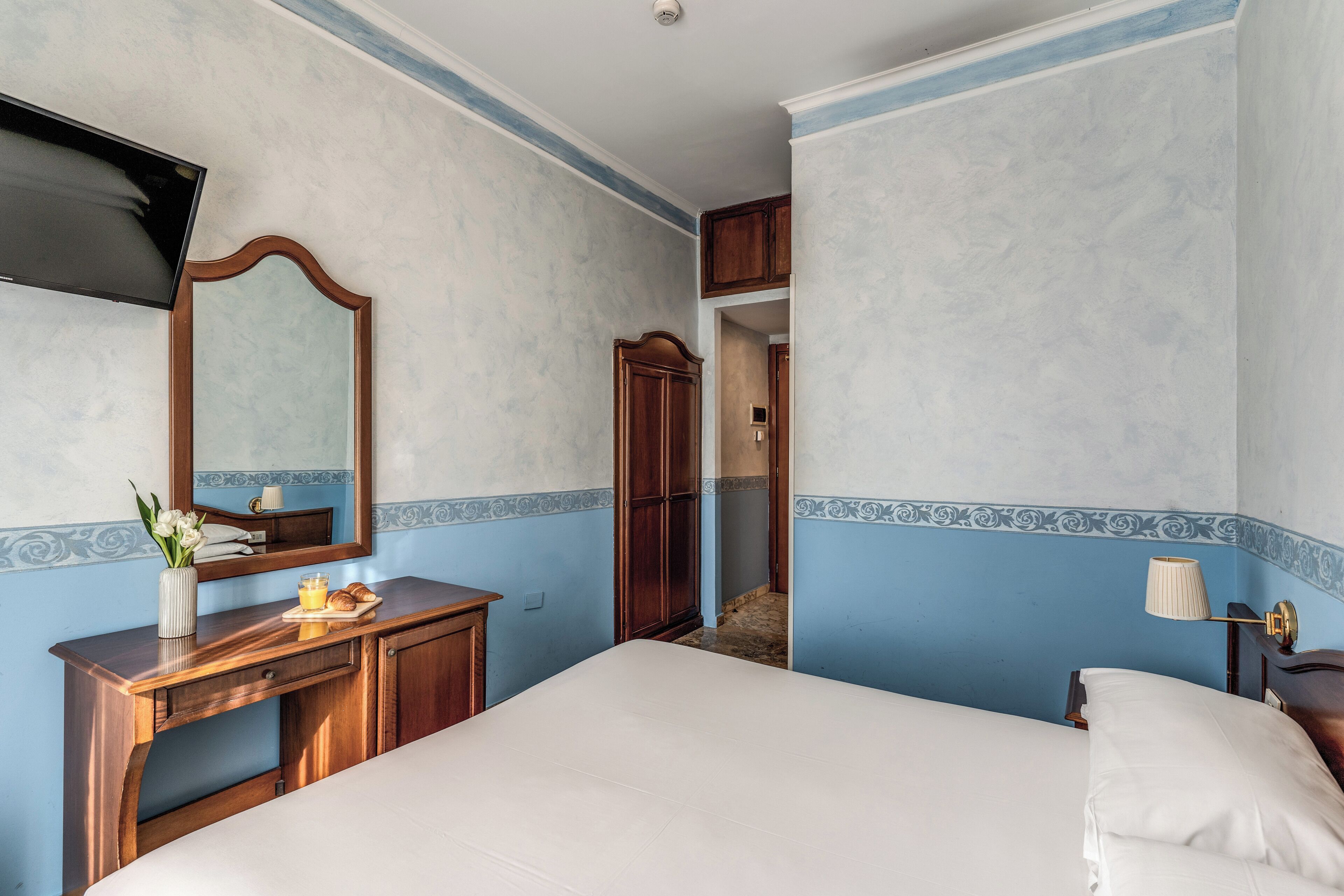 A hotel room with a bed, a wooden dresser, a mirror, and a nightstand with a lamp.