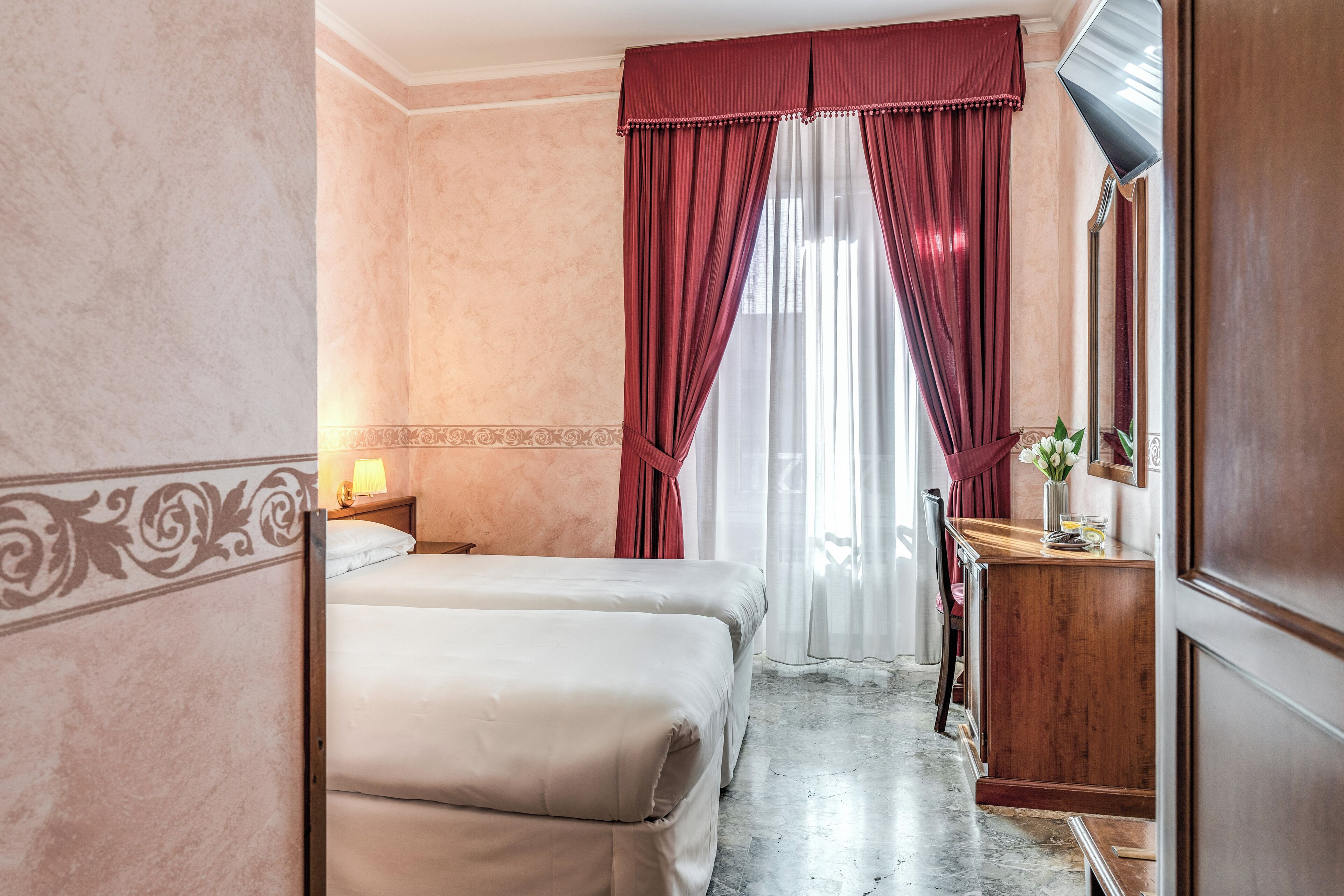 Habitación de hotel con dos camas, un aparador de madera, un espejo y una ventana con cortinas rojas.