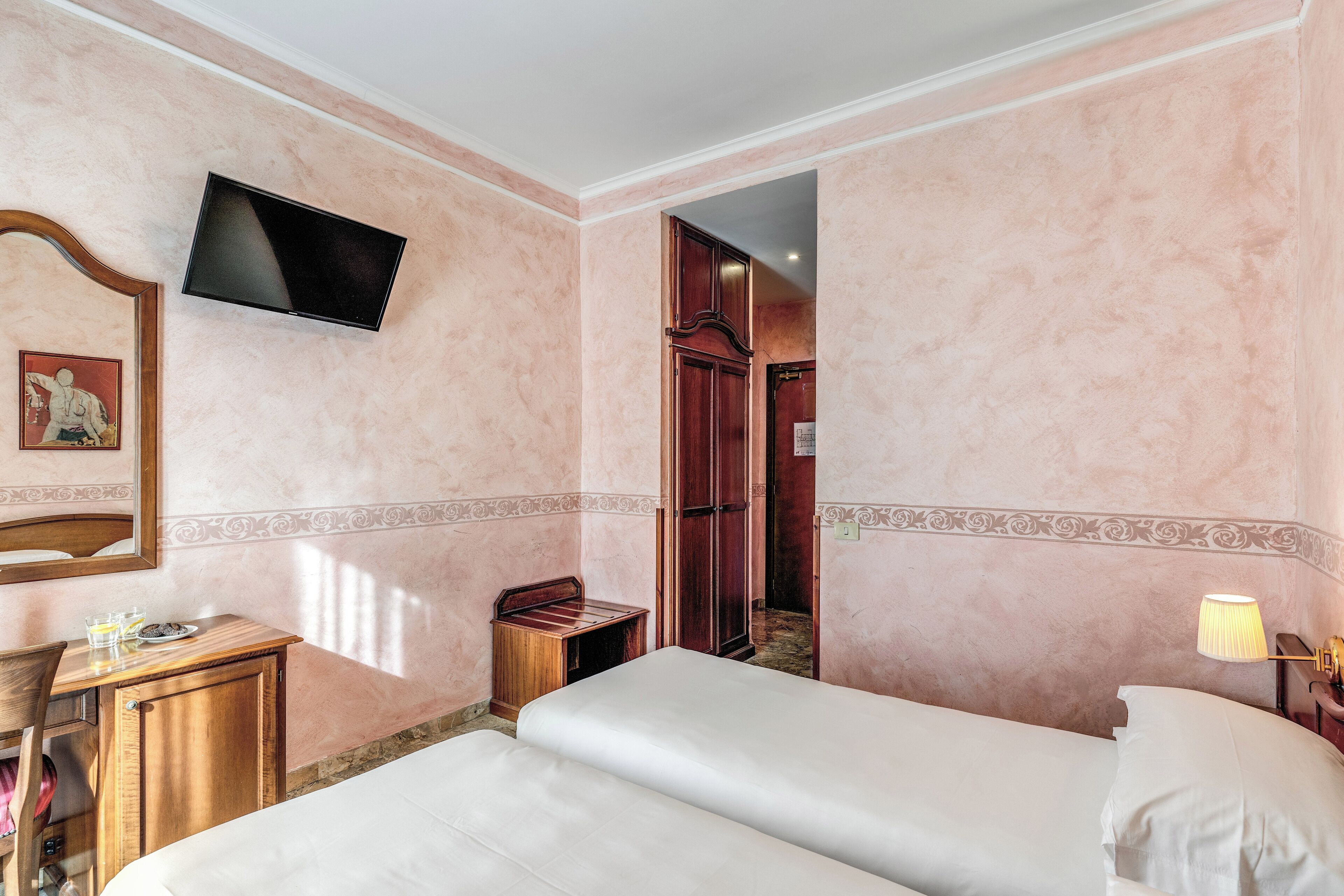 A hotel room with two beds, a TV mounted on the wall, a desk with a chair, and a wooden cabinet.