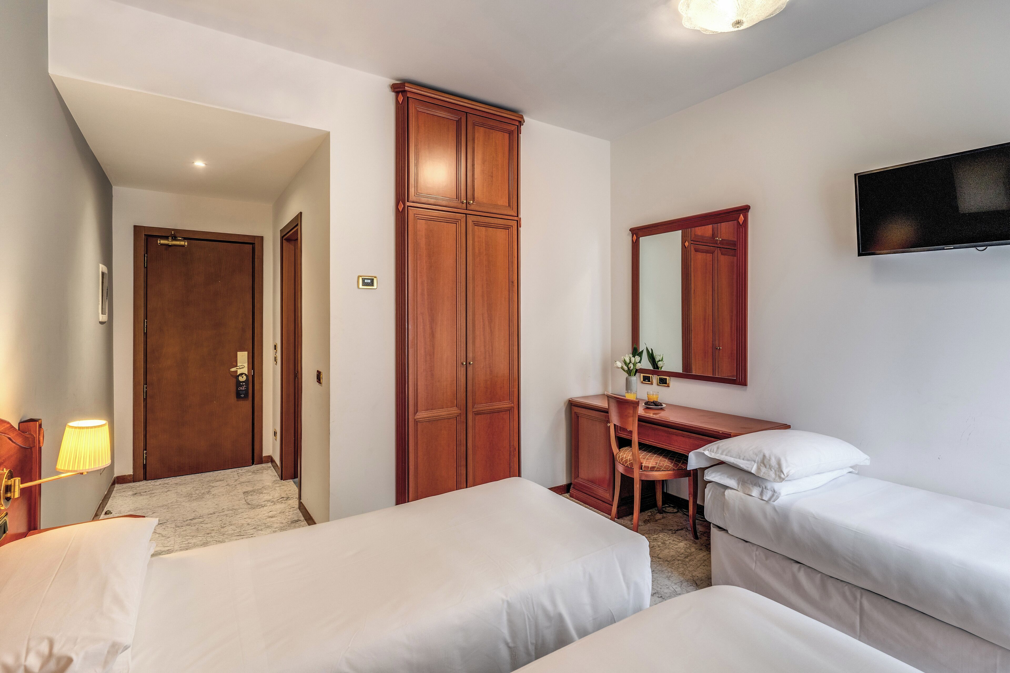A hotel room with two single beds, a wooden wardrobe, a desk with a chair, a mirror, and a television mounted on the wall.