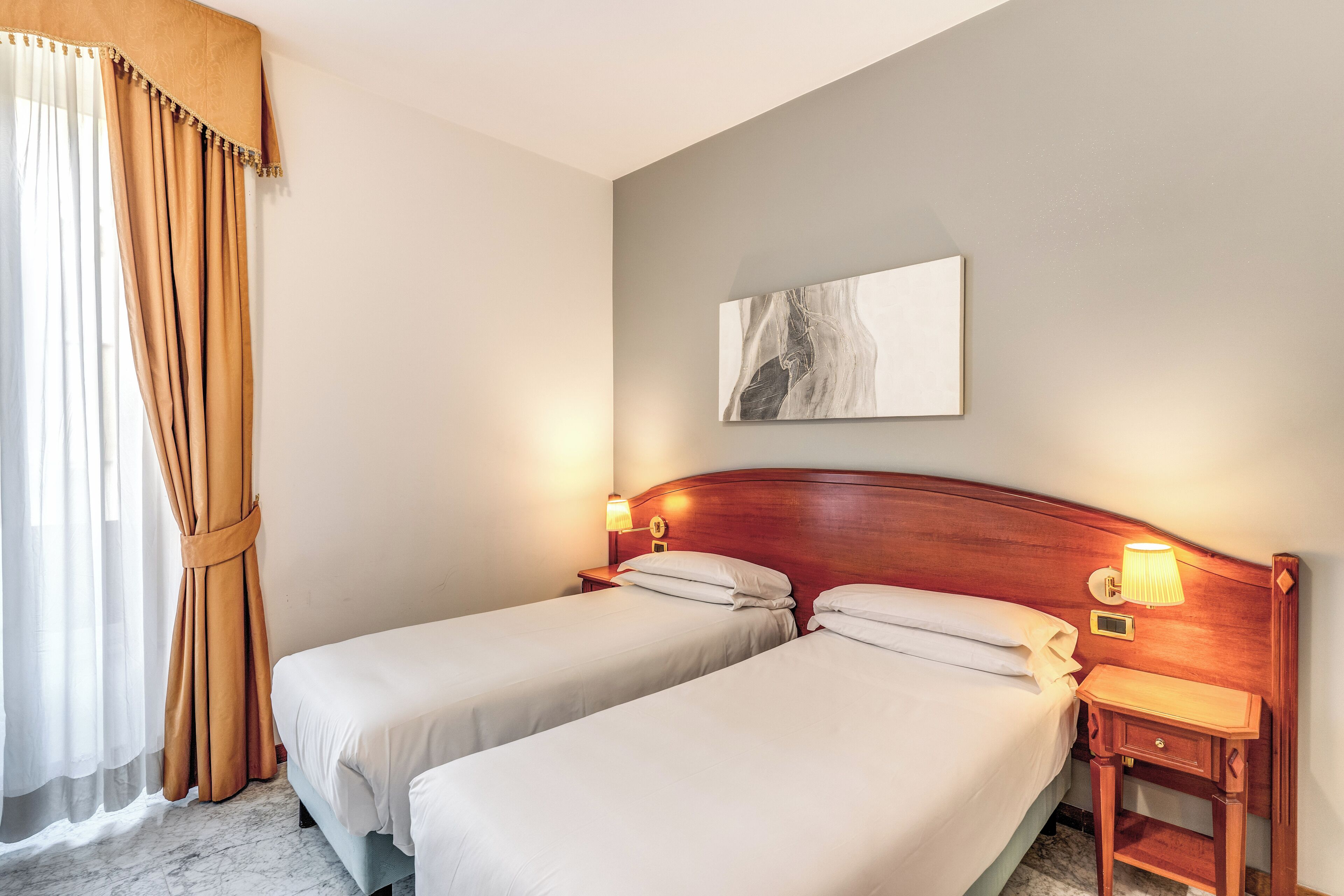A hotel room with two single beds, a wooden headboard, bedside lamps, a nightstand, a marble floor, and a large abstract painting on the wall.